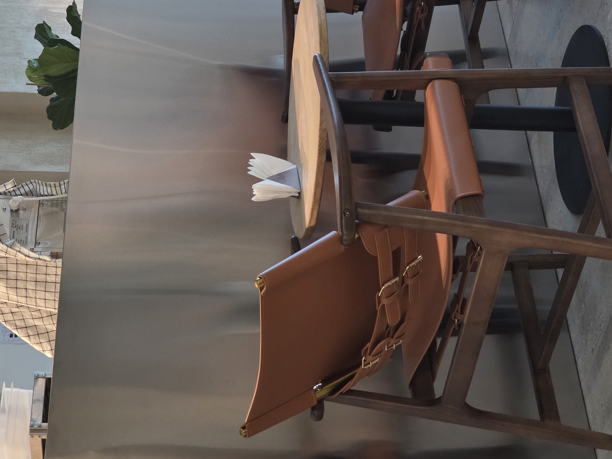 This image depicts a cafe or restaurant setting in an unknown city. A stainless steel table is flanked by chairs featuring wooden frames and tan leather seats and backs. A stack of paper napkins sits on the table. A potted plant adds a touch of greenery to the space. The floor appears to be made of concrete.