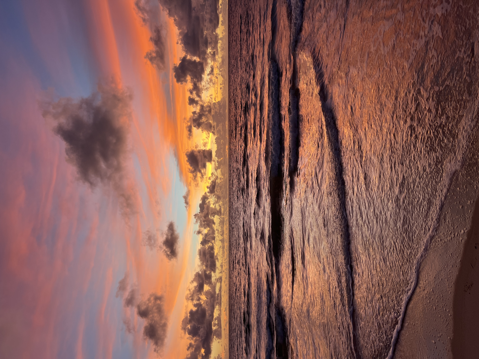 The image depicts a serene beach scene at sunset. The composition is split vertically, with the left half showcasing a dramatic sky filled with swirling clouds painted in hues of orange, pink, and purple, transitioning to a soft blue at the top. Darker, ominous clouds are scattered amongst the vibrant colors, creating a striking contrast.
The right half of the image is dominated by the ocean and the shoreline. Gentle waves are captured in motion, reflecting the warm, golden light of the setting sun. The water's surface is textured, with clear lines of crests and troughs. The wet sand of the beach is visible in the bottom right corner, showing subtle ripples left by the receding water.
The time of day is clearly sunset, indicated by the intense colors in the sky and the warm lighting on the water and sand. The weather appears calm, with soft light and gentle waves. No people or man-made objects are visible, emphasizing the natural beauty of the location. The overall mood is tranquil and awe-inspiring, evoking a sense of peace and the grandeur of nature. The location is a beach, but specific geographical cues are absent. There is no visible text in the image.
