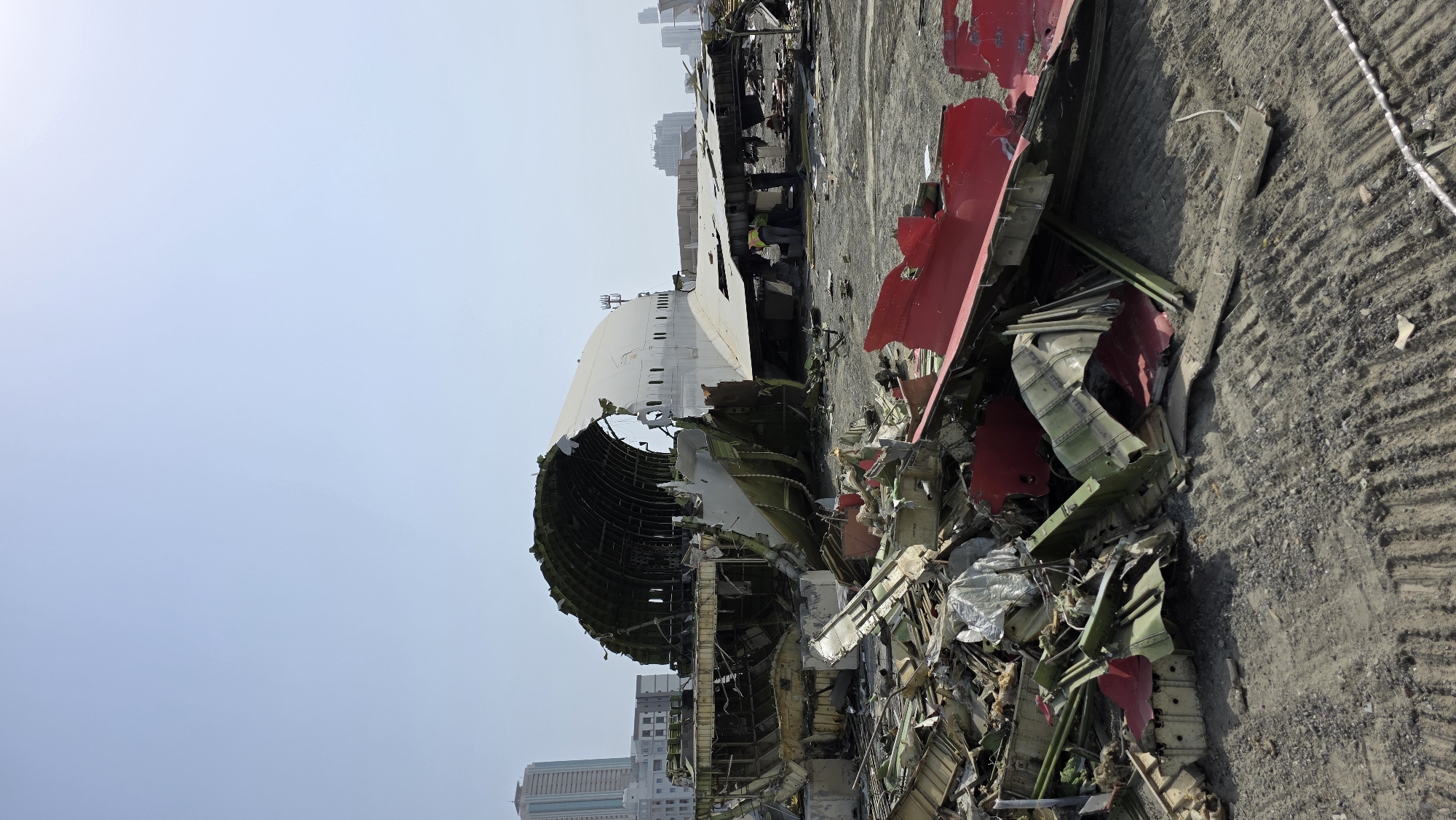 The image depicts the wreckage of a large aircraft, possibly a Boeing 747, scattered across a dry, dusty lot in Fujairah, United Arab Emirates. The main fuselage section is heavily damaged, with a large portion of the upper hull torn away, revealing the internal structural framework. Sections of the plane's exterior, including large white and red panels, are strewn about the foreground and midground, intermingled with debris. Several figures, presumably workers wearing high-visibility vests, are visible near the wreckage, suggesting ongoing salvage or dismantling operations. In the background, modern high-rise buildings of Fujairah can be seen against a clear, pale blue sky, providing a stark contrast between the urban development and the scene of destruction. The ground is a mixture of dirt and gravel, marked with tire tracks and footprints.