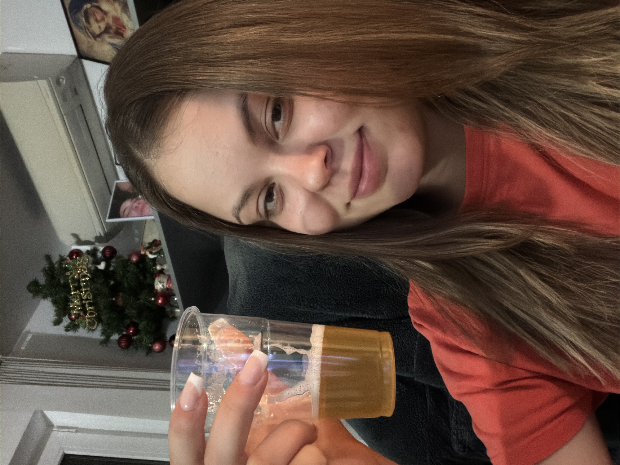 A young woman is holding a clear plastic cup with an orange beverage in it, possibly beer, up to the camera. She is smiling and looking at the camera. She has long, light brown hair and is wearing an orange shirt. In the background, there is a small Christmas tree with red ornaments and a gold "Merry Christmas" sign. There is also a white cabinet and what appears to be a framed photo of a person or people. Given the presence of the Christmas tree, it is likely that this photo was taken during the Christmas season. There is no way to determine the location is Varna, Bulgaria from the given information.