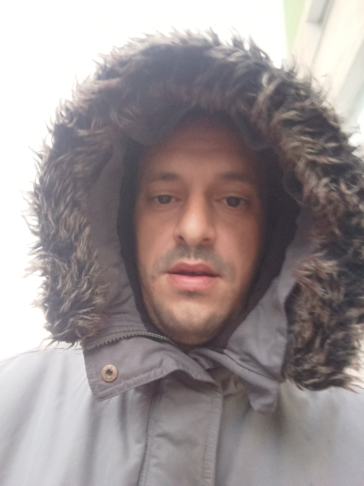 A close-up shot captures a middle-aged man with light skin under a hooded winter coat with fur trim. His face is framed by the hood, and he has short, dark hair and a light stubble. The coat is a light grey color. The man is likely outdoors. There is nothing in the image to indicate the location is Burgas, Bulgaria.