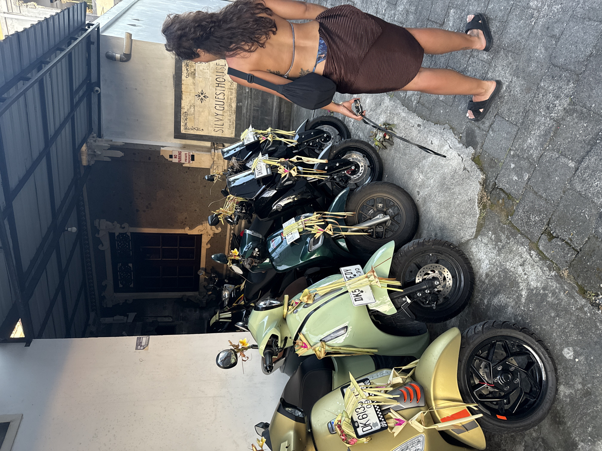 The image captures a partial view of a street in Canggu, Indonesia. The main subject is a row of parked motorbikes, adorned with traditional Balinese offerings, including woven palm leaves and small flowers. The license plates of some of the motorbikes are visible, including "DK 613," "DK5 FDY," and "DK6127." A sign reading "SILVY GUESTHOUSE" and "NO PARKING PRIVATE PROPERTY" is visible on the wall behind the motorbikes. A woman wearing a brown skirt and sandals is positioned to the side, holding a device. Her back is turned to the camera, and she appears to be observing the motorbikes. The environment includes a paved street and a building with an overhanging roof.