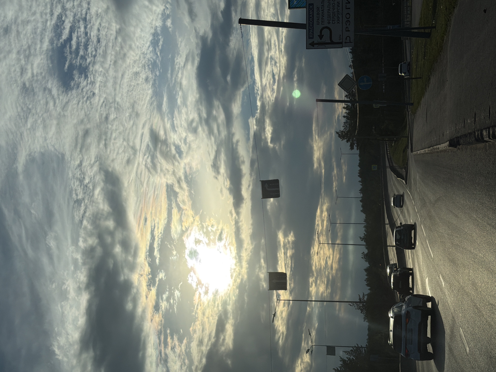This is a photograph taken from a vehicle, looking towards the sky and the side of a road. The scene depicts a road with several cars traveling in the same direction. The sky is filled with dramatic clouds, with the sun shining brightly through them, creating a hazy and somewhat iridescent effect. The time of day appears to be late afternoon or early evening, given the angle of the sun and the long shadows cast on the road. The weather is overcast but with breaks of sunlight.
On the left side of the road, there are several tall poles with lights, suggesting a highway or a well-lit road. There are also signs visible, one of which has text in Russian. The text on the most prominent sign reads "КРАСНОКАМСК" (Krasnokamsk) and also shows directions with an arrow pointing left and another indicating a turn. Below this, there are words like "КРАЕВОЙ", "ПЕРИНАТАЛЬНЫЙ", "ФЕДЕРАЛЬНЫЙ", "СЕРДЕЧНО-СОСУДИСТОЙ", and "ХИРУРГИИ". Another sign further down the road appears to have "РЭО ГИ" and "50км". These are likely signs related to a city or region and distances to medical facilities.
The overall mood of the image is atmospheric and dynamic, due to the interplay of light and shadow in the clouds and on the road. The presence of the cars indicates a travel or commuting scenario. The setting appears to be a road leading out of or towards Krasnokamsk, Russia.
