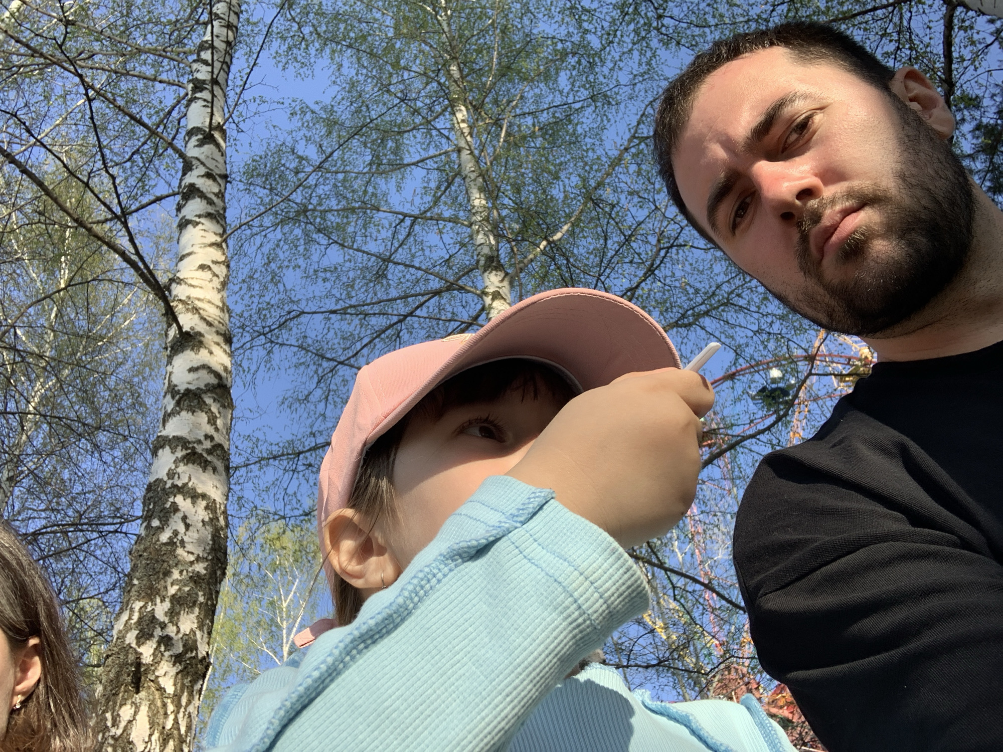 The image is a low-angle shot capturing two individuals and a partial third person outdoors, likely a selfie or taken by someone nearby. A bearded man with dark hair occupies the upper right portion of the frame, wearing a black shirt. A child with a pink cap is positioned slightly lower, wearing a light blue sweater. The child is holding something white near their mouth, obscuring part of their face. Part of a woman's face appears on the bottom left edge. The background shows birch trees with bare branches against a blue sky. A Ferris wheel is visible through the trees. The location is Chaikovsky, Russia.