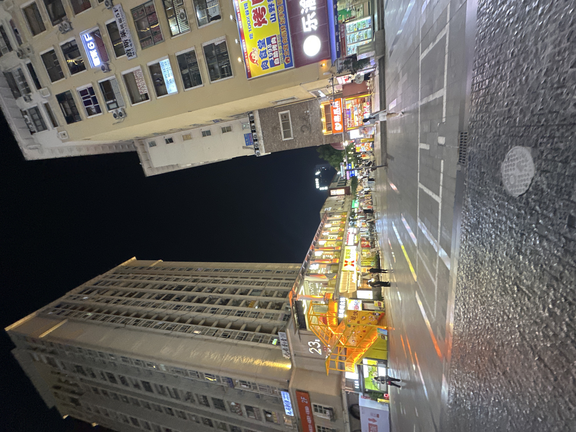 The image depicts a lively pedestrian street scene at night in the Chongqing High-tech Zone, China. Two tall buildings, one on the left and another on the right, flank a wide, wet street. The building on the left is a light-colored high-rise with many windows. The building on the right is a shorter, yellow-toned building with multiple levels of windows and prominent signage, including a bright "6F" sign and other advertisements.
In the foreground, the street surface is a mixture of grey cobblestones and paved sections with painted white lines, suggesting a crosswalk or lane markings. The wetness of the pavement indicates recent rain or cleaning, reflecting the colorful lights from the surrounding buildings.
Along the right side of the street, a series of shops and restaurants are visible, illuminated by neon signs and interior lighting. These establishments feature awnings and outdoor seating areas. People are scattered throughout the scene, walking, standing, and interacting. One person is walking away from the camera on the right side of the street, while others are visible in the middle ground, browsing shops or conversing.
A distinctive bright orange and yellow staircase is a prominent feature on the left side of the street, leading up to a multi-level structure that appears to be part of the shops or restaurants. The number "23" is clearly displayed near the base of this staircase.
The overall atmosphere is one of urban nighttime activity, with vibrant lights, wet surfaces, and the presence of pedestrians suggesting a bustling commercial and social area. The dark night sky provides a backdrop against which the illuminated buildings and signs stand out sharply.