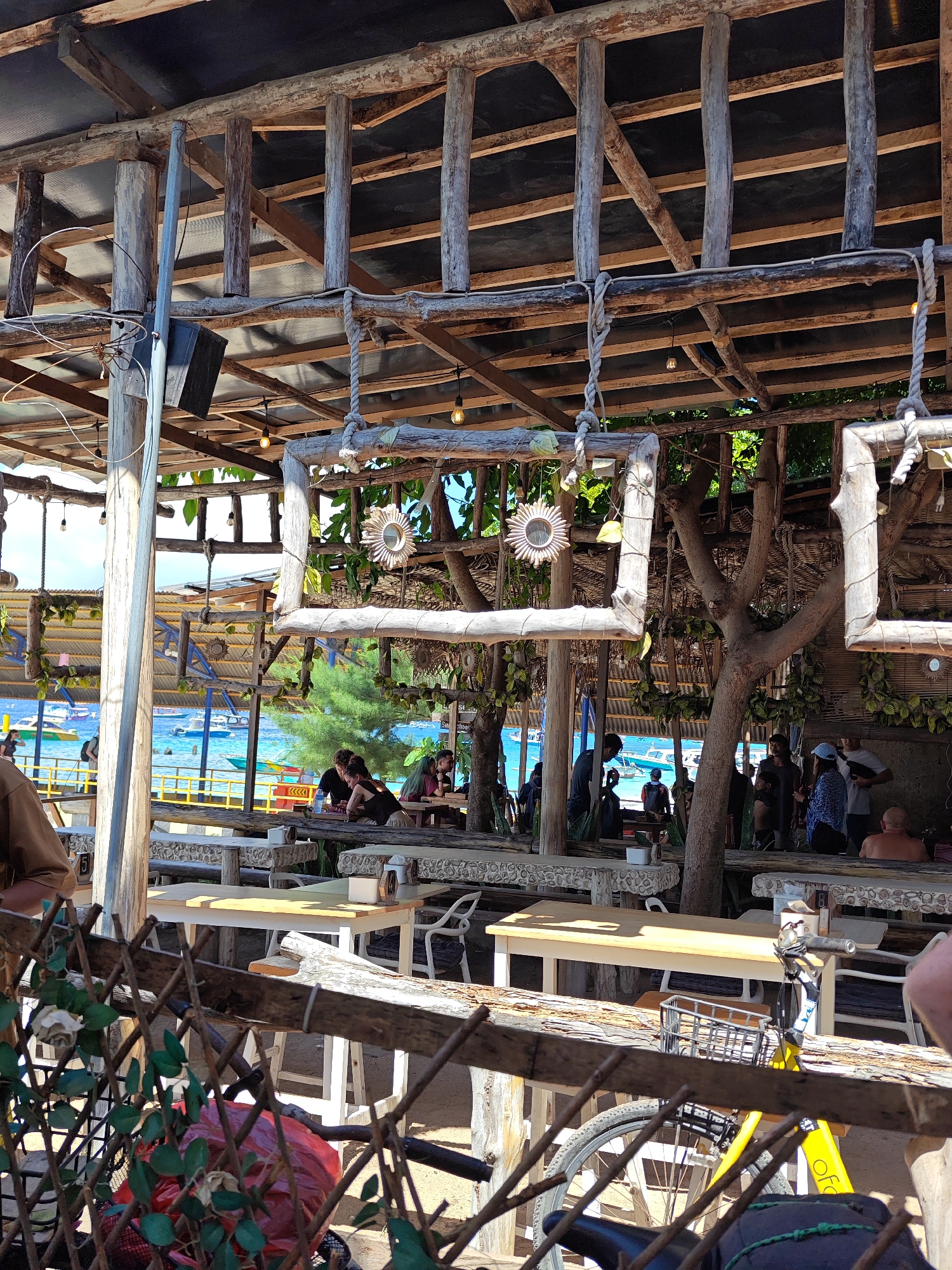 The image depicts a restaurant scene on a beach in an unknown city in Indonesia. The restaurant features rustic wooden structures with open walls providing views of the sea and boats. Several people are seated at tables, appearing to be dining or socializing. Others are standing or walking nearby. Decorative elements include wooden frames hanging from the ceiling. A fence partially obscures the foreground, with foliage and a bicycle visible. The overall atmosphere suggests a relaxed, seaside setting.