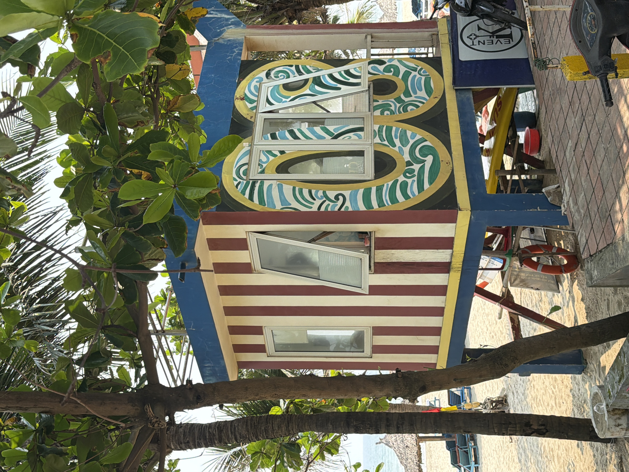 The image shows a small, elevated structure, possibly a lifeguard station or small beachside business, located in Đà Nẵng, Vietnam. The structure has two levels. The upper level has a painted design featuring a figure-eight pattern with blue, green, and gold colors. The lower level has horizontal stripes of red and white. Both levels have windows, some of which are open. The structure is elevated on supports painted blue. A motorcycle is parked nearby. A life preserver hangs on the side of the structure. A tree with green foliage obscures part of the view. The ground appears to be a combination of sand and paved walkway.