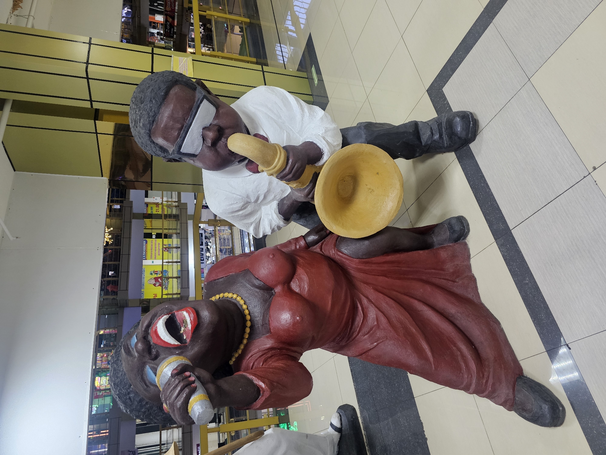 Two life-size statues of Black musicians are depicted in a public indoor space, likely a shopping mall, with a checkered tiled floor and prominent yellow and white structural columns in the background.
On the left is a female statue of a singer, rendered in a dark brown tone. She is depicted in a dynamic pose, leaning forward with her mouth wide open as if singing into a silver microphone she holds in her right hand. She wears a long, form-fitting red dress, a yellow beaded necklace, and her eyes are painted blue. Her body is exaggerated in form.
To the right of the singer stands a male statue, also in a dark brown tone. He is dressed in a white collared shirt, dark pants, and wears thick-rimmed silver glasses. He is playing a yellow saxophone. His body is also rendered with exaggerated features, and he appears to be slightly hunched over his instrument.
The background shows the interior of a commercial building, with commercial signage and lighting visible on multiple levels. The overall impression is a festive and artistic display, possibly celebrating music or culture. The location is specified as the city of Nakhodka, Russia.