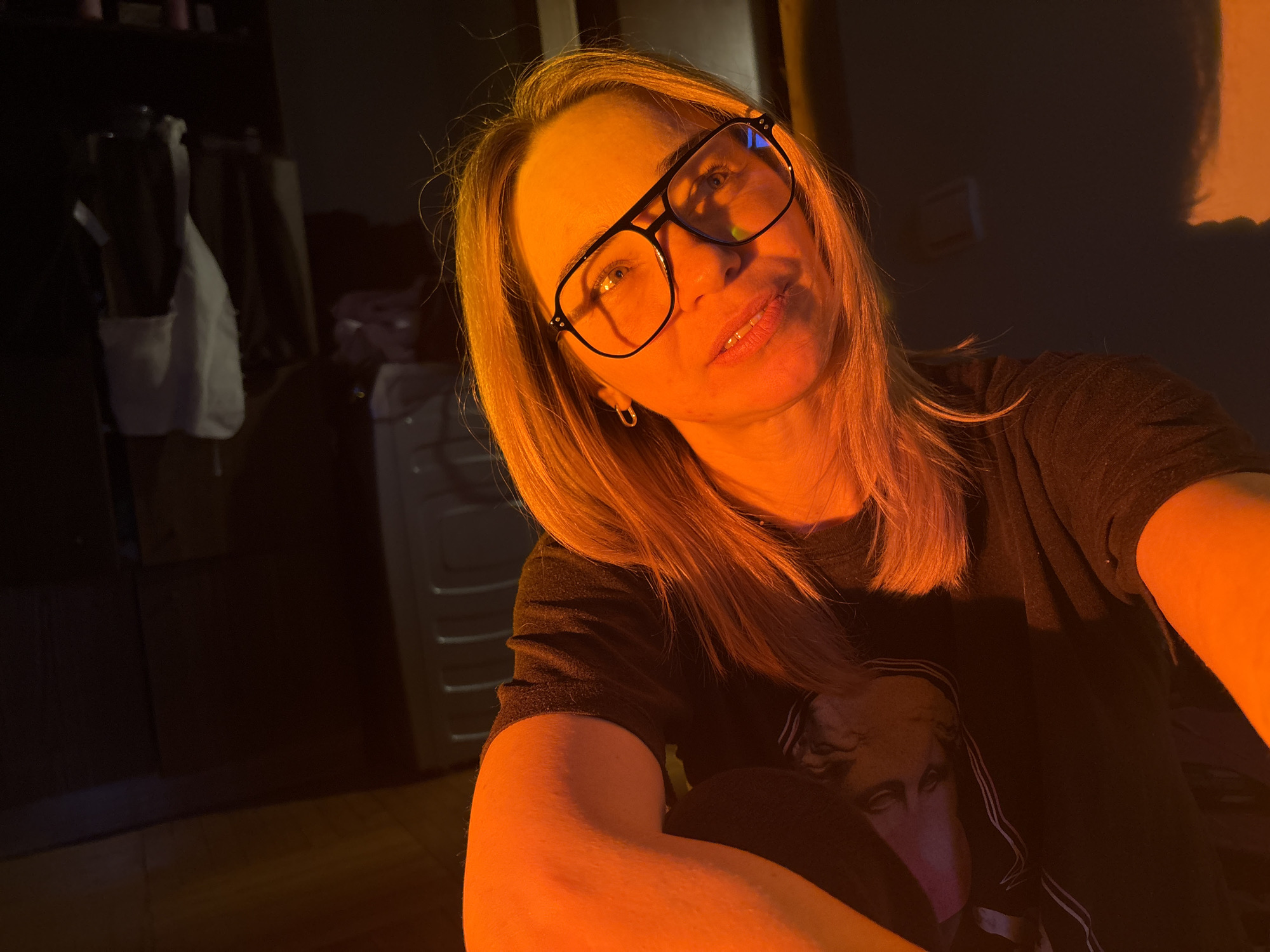 A woman with light-colored hair and black-framed glasses is captured in a close-up, likely a selfie, in a dimly lit indoor setting in Russia. Her face is softly smiling and directly facing the camera, illuminated by a strong, warm orange light that casts deep shadows elsewhere. The orange light highlights her hair and skin, creating an intimate and cozy atmosphere.
She is wearing a dark t-shirt with a graphic print featuring an indistinct artistic depiction of a face, possibly a classical bust or a stylized portrait. The background is largely dark, suggesting night or a low-light environment. To the left, dark furniture and a white hanging object, possibly a garment or bag, are barely discernible. On the far right, a wall reflects the same orange glow, further enhancing the warm lighting effect. No specific text is visible in the image.