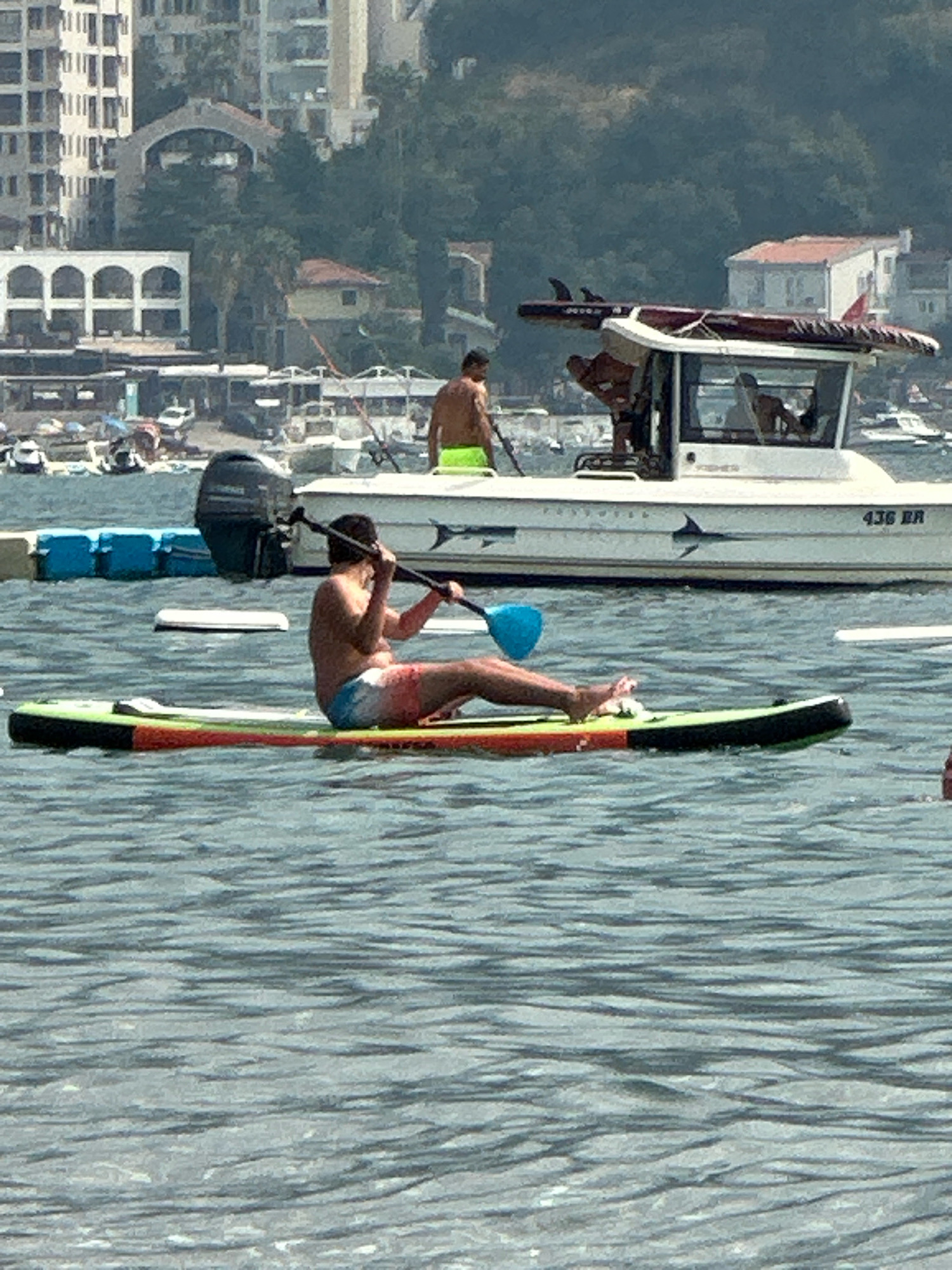 The image captures a sunny day in the city of Boreti, Montenegro. In the foreground, a person is paddleboarding on the water, holding a paddle with a blue blade. The paddleboard is multi-colored, with green, orange, and red accents. The person is wearing patterned shorts. In the background, a white boat with a cabin is visible, with surfboards on its roof. Another person, wearing bright green shorts, is standing on the boat. Beyond the boat, buildings and a forested hillside are visible, suggesting a coastal setting. The water appears calm with ripples.