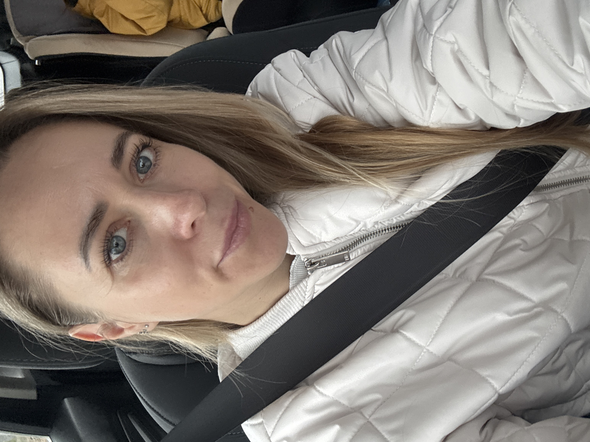 The image shows a woman with light blonde hair and blue eyes wearing a white quilted jacket and a seatbelt, taken from a low angle. She is likely in a car, based on the presence of the seatbelt and what appears to be a car seat visible in the periphery. There is no information in the image to indicate that the location is Chaikovsky, Russia. The environment is not detailed enough to determine a specific location.