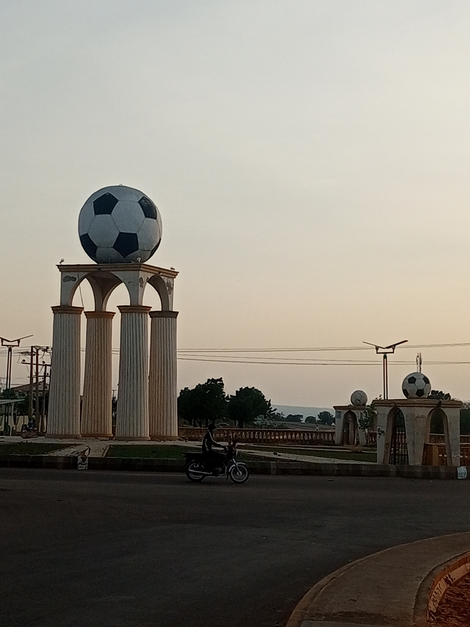 The image shows a roundabout in the city of Gombe, Nigeria. A large, black and white soccer ball sculpture sits atop a monument with four white pillars and an arched structure. In the middle ground, a person rides a motorcycle on the road. Several trees are visible in the background, along with a faint outline of hills. The sky is overcast, suggesting it is either early morning or late afternoon. The overall scene depicts a public space with a prominent decorative element.