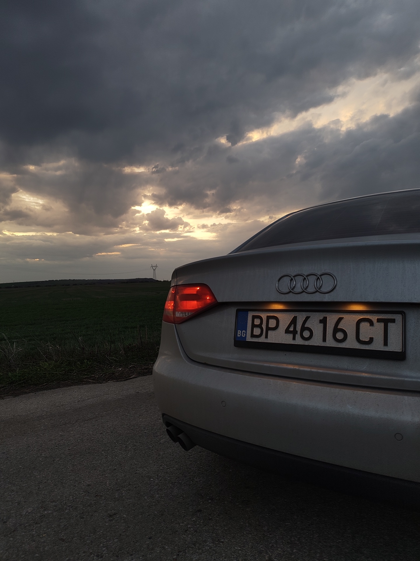 A silver Audi sedan, displaying Bulgarian license plate 'BP 4616 CT', is parked on the asphalt shoulder of a road in Gorni Tsibar, Bulgaria. The vehicle is positioned facing away from the viewer, with its rear-left quarter visible. The driver-side taillight is illuminated red, and the license plate lights are active, casting an amber glow. Dual exhaust pipes are visible on the lower left rear, and circular parking sensors are embedded in the bumper. To the left of the road, a green field extends into the distance. A tall, lattice-style metal power line tower is visible on the horizon, silhouetted against the sky. The sky is dominated by dark, heavy grey clouds, through which distinct patches of golden-orange light are breaking, suggesting either sunset or sunrise. No individuals or direct actions are observable.