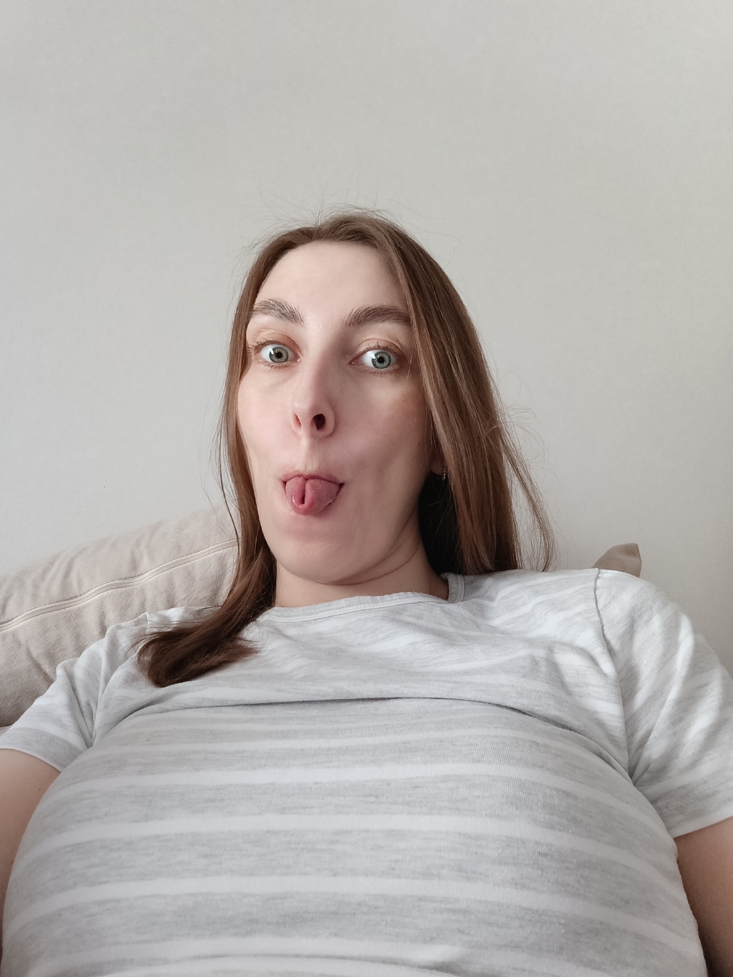 A close-up, slightly low-angle photograph features an adult female indoors, striking a playful pose. She has long, straight brown hair, fair skin, and wide, bright green or blue eyes. Her eyebrows are prominent. She is making a humorous facial expression, sticking out her tongue, which is slightly curled, and her mouth is open.
She is wearing a casual, light grey t-shirt adorned with thin, horizontal white stripes. To her left and behind her, a portion of a light beige or off-white cushion or pillow is visible, suggesting she is seated or reclining on a piece of furniture like a sofa or bed. The background is a plain, light-colored wall, indicating an indoor setting, likely a home. The lighting is soft and even, consistent with natural indoor ambient light during the day.
The image conveys a sense of humor and lightheartedness. No text is visible within the frame. The contextual location is Chaikovsky, Russia.