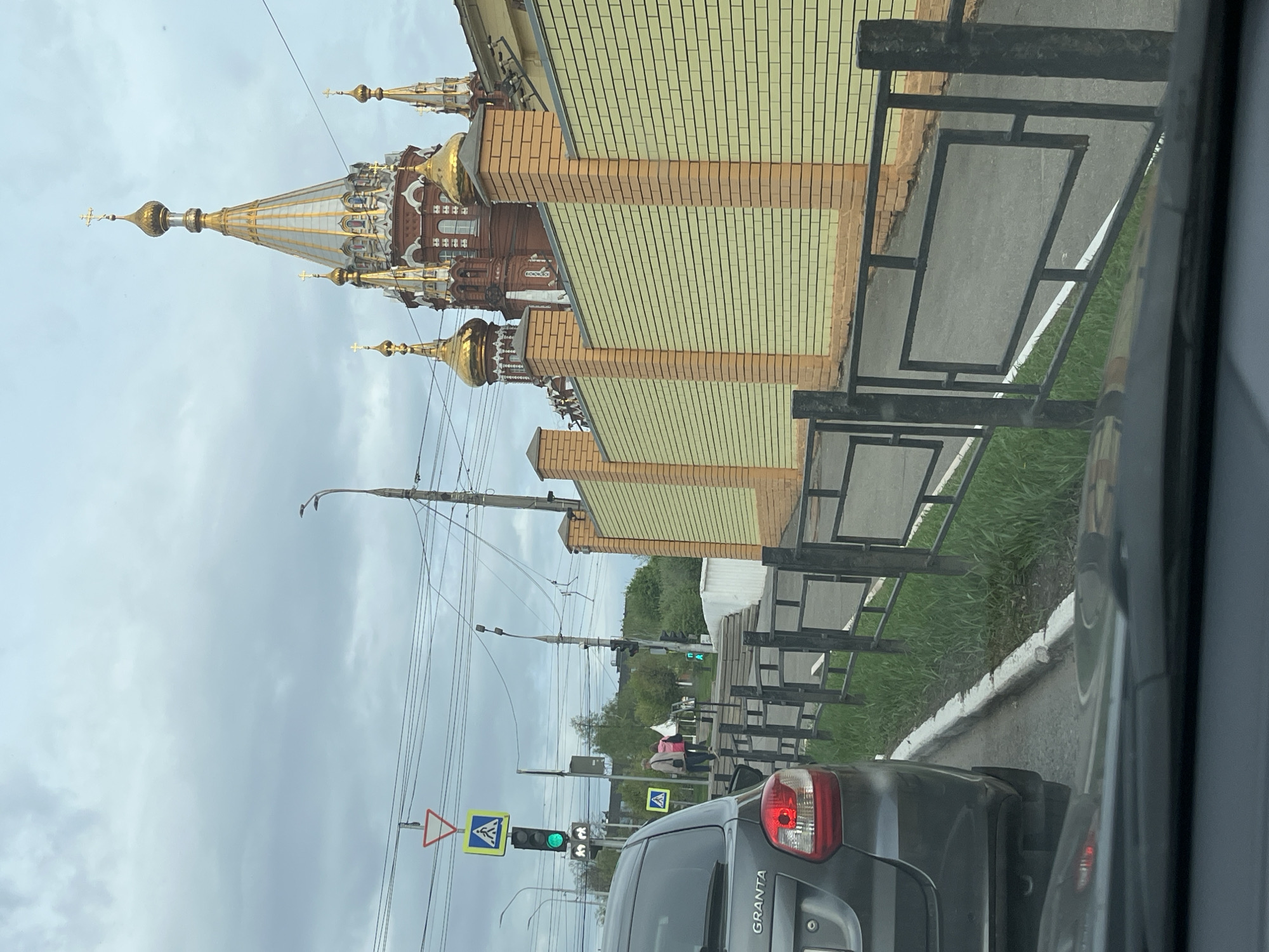 The image, taken from inside a vehicle, shows a street scene in Izhevsk, Russia, featuring the Saint Michael Cathedral. The cathedral's distinctive golden domes and ornate spires are visible against a cloudy sky. A building with brick and yellow tile facade runs along the side of the street. Utility poles and wires stretch across the scene. The rear of a dark grey "Granta" car is visible in the foreground, with a traffic light showing a green light. Pedestrian crossing signs are present, and indistinct figures of people are seen crossing the street. A metal railing lines the sidewalk.
