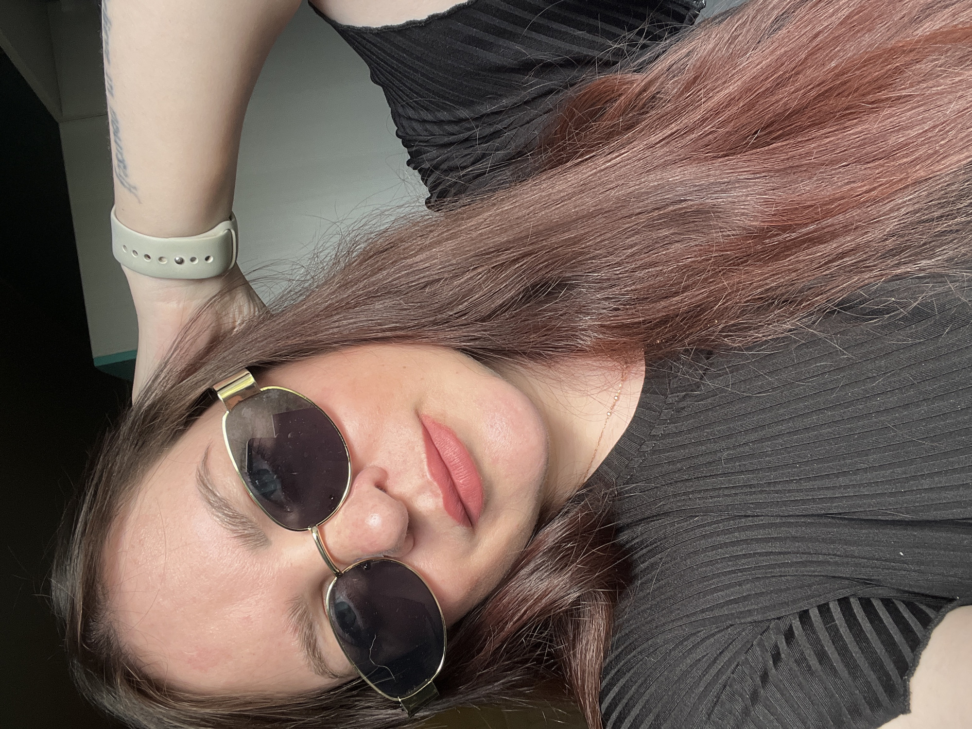 A woman with long, reddish-brown hair is wearing dark sunglasses and a black ribbed top. She has a light-colored watch on her left wrist. The background appears to be a simple interior space. The location is unspecified and unable to be determined from the image.