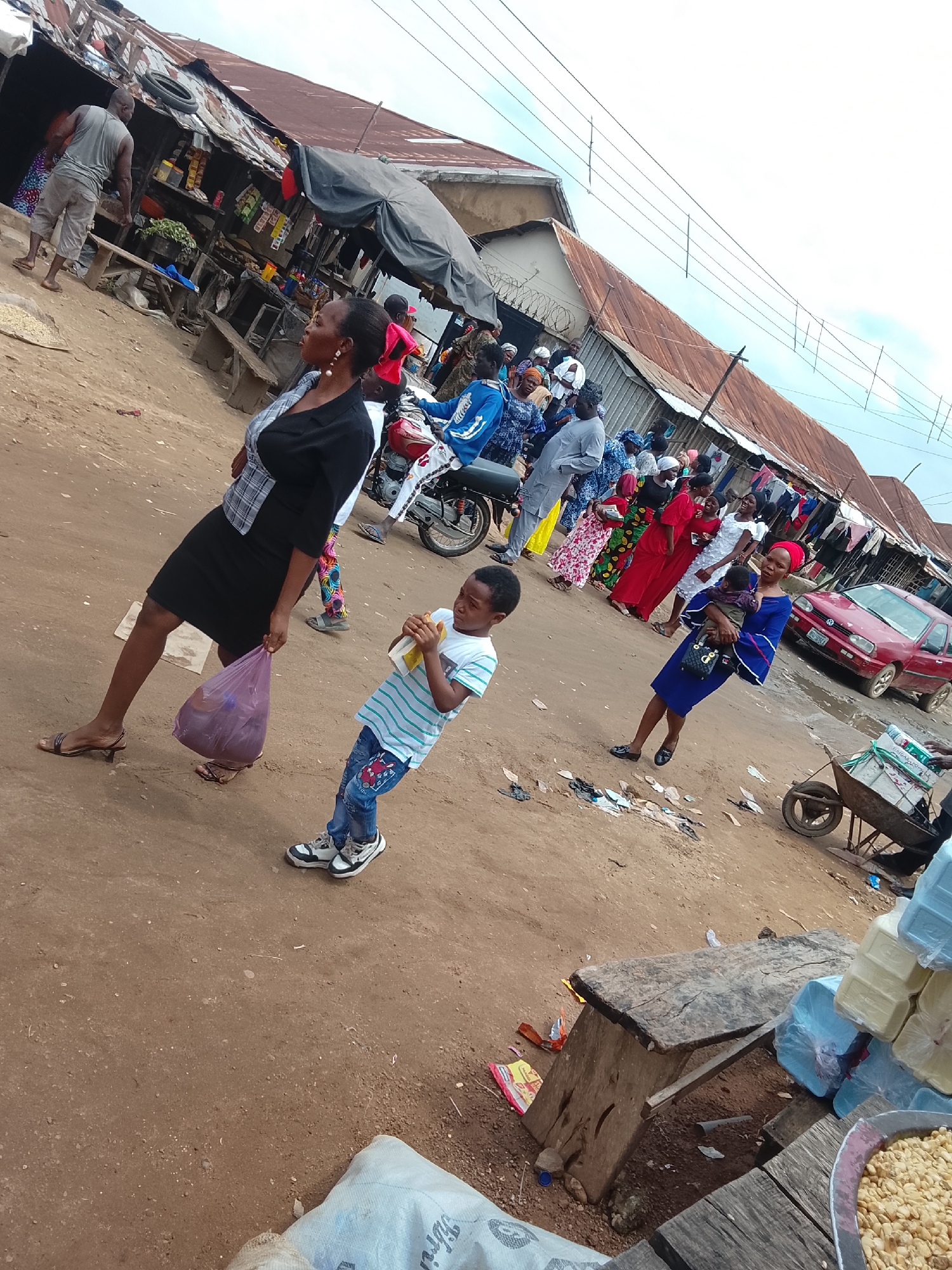 This image captures a bustling market scene in Abuja, Nigeria, on a cloudy day. In the foreground, a woman in a black dress and a checkered top walks away from the camera, carrying a purple plastic bag. To her right, a young boy in a striped t-shirt and jeans stands holding something yellow and white, looking towards the left. Behind them, a man in a grey tank top and shorts is visible near a market stall made of corrugated metal and wood, with various goods displayed on shelves. Several other people are scattered throughout the scene, some appearing to be shoppers and vendors. A red car is parked on the right side of the frame, and a wheelbarrow loaded with white packages is also present. The ground is unpaved dirt, and litter is visible in places. The overall atmosphere suggests a busy local marketplace.