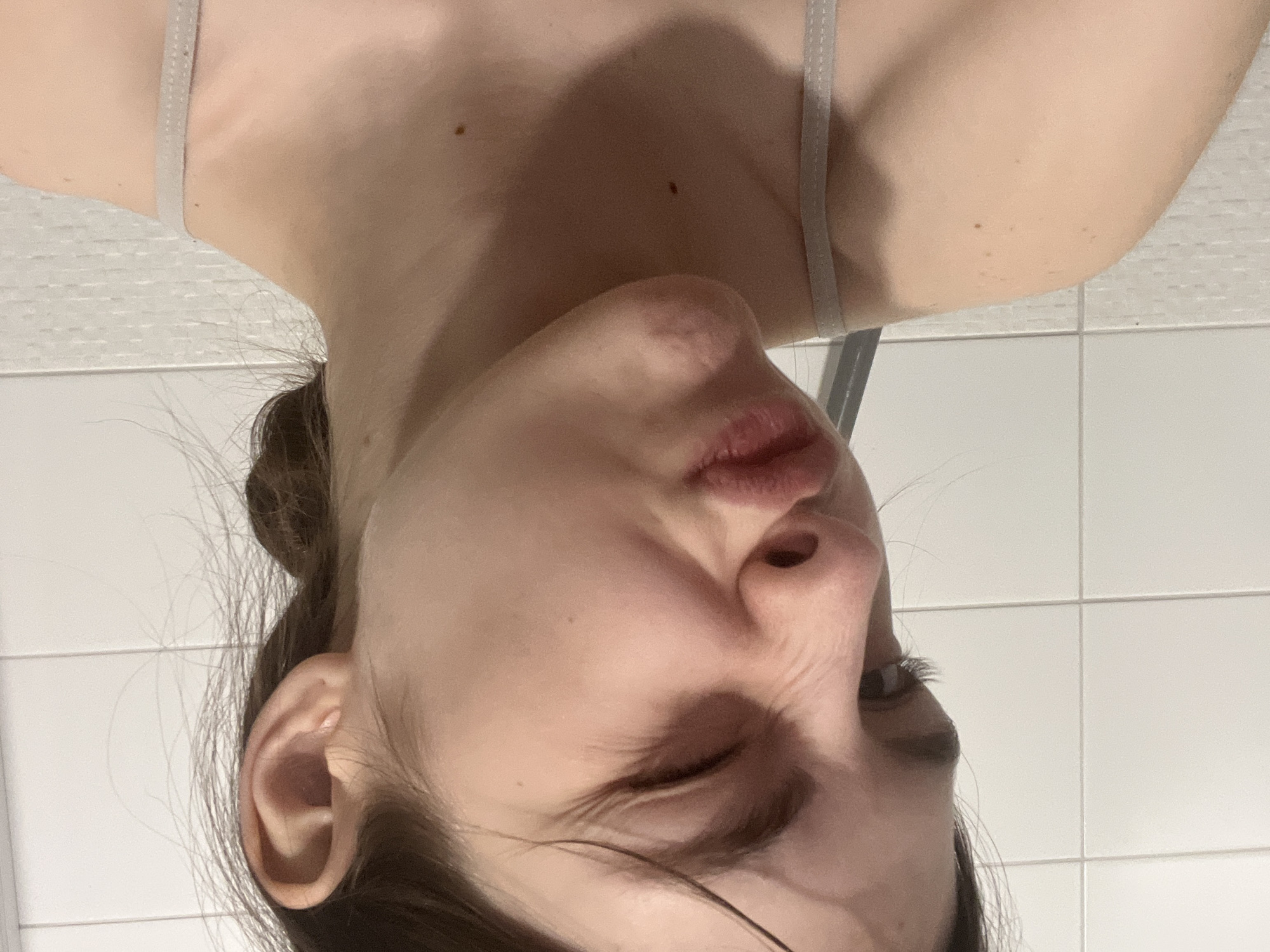 A close-up, inverted image of a person's face. The person is fair-skinned with brown hair pulled back. One eye is closed or squinted. The person's lips are pursed, and the face appears to be in a state of contortion or grimace. The background shows white tiled walls. There is no indication of this being located in Saint Petersburg, Russia.