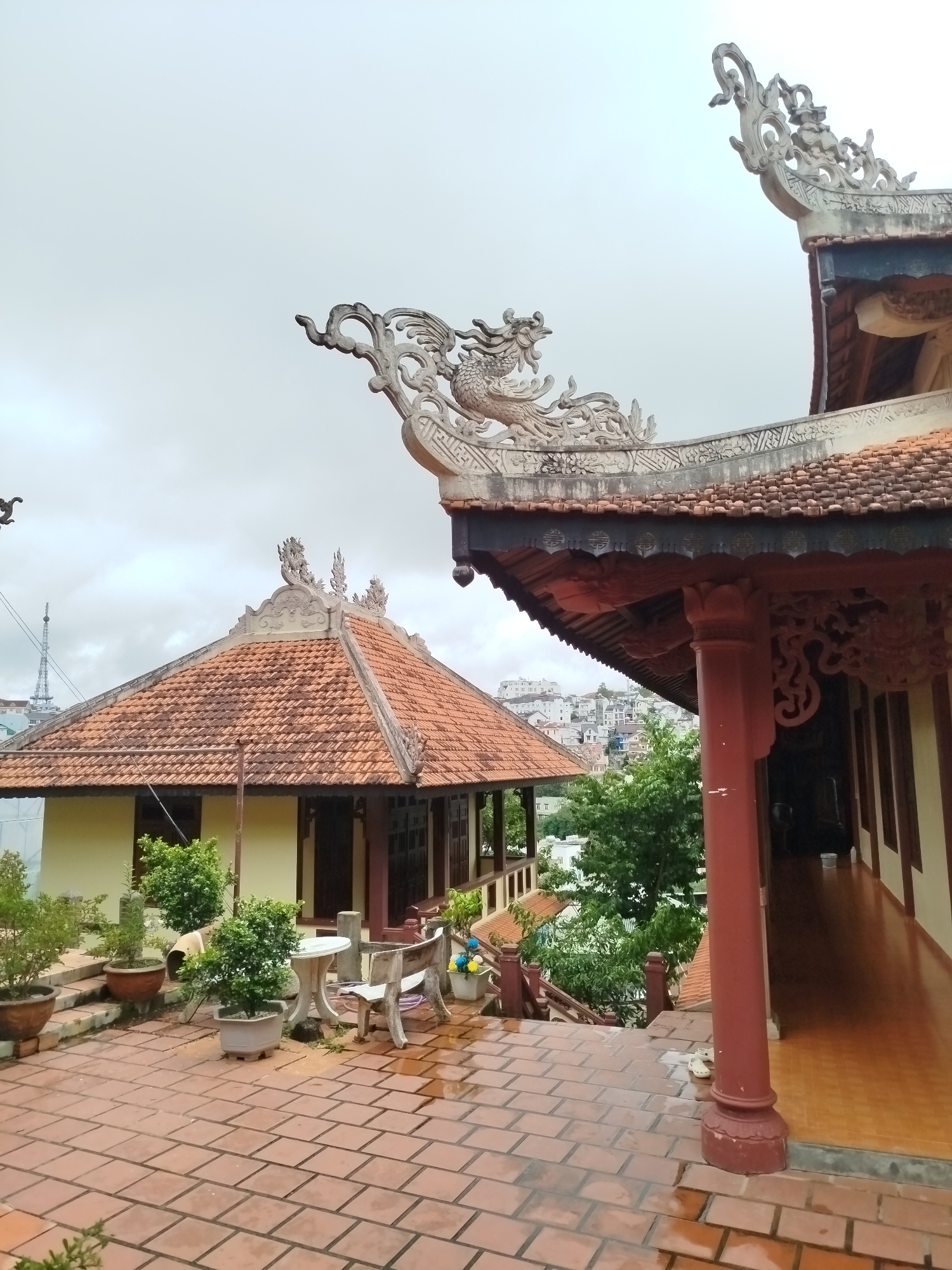 This is a medium shot, eye-level, exterior photograph of a Buddhist temple in Da Lat, Vietnam, on an overcast day. The foreground is a red-tiled patio, with some areas appearing wet. To the left, a light yellow building with a red-tiled roof has several potted plants on its patio. In front of this building, a white table and two chairs are visible, along with a wooden bench. A walkway with red-tiled flooring and orange pillars runs along the right side of the image, leading to a covered porch with ornate carvings. Two buildings with traditional Vietnamese architecture are prominently featured, with decorative dragon motifs on their roofs. In the background, a city with houses and green trees is visible on a hill. A tall tower with a radio antenna is also present in the distance.