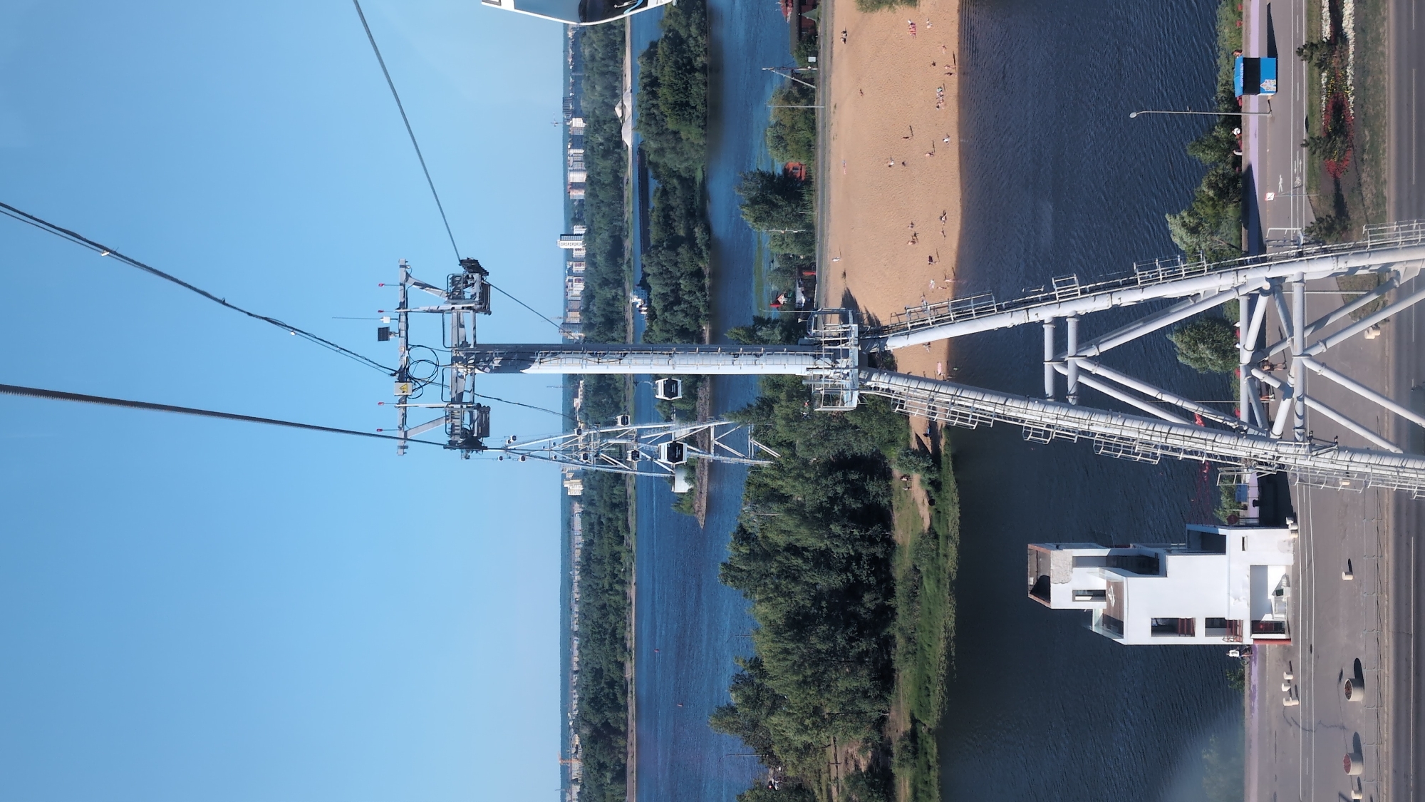 This is an aerial, vertically oriented, medium shot of a cable car system over a river in Nizhny Novgorod, Russia. The sky is clear and blue, suggesting a sunny day. The cable car's support structure, made of white metal, dominates the frame, with multiple cable cars visible suspended from its cables. Below the cable car system, a wide river flows, bordered by lush green trees on one bank and a sandy beach on the other. Several people are scattered on the beach, enjoying the sunny weather. Across the river, a road runs alongside the beach, with a few cars and buildings visible in the distance. A modern white building with large windows is situated near the riverbank, below the cable car's support structure. The scene appears to be a recreational area, likely a park or a popular spot for tourists and locals. The overall impression is one of a vibrant, outdoor activity on a pleasant summer day.