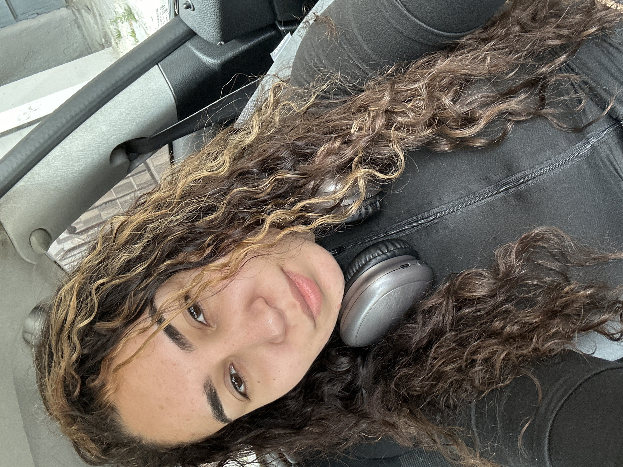 A woman with curly brown and blonde hair is reclining in the passenger seat of a car. She is wearing a black zippered top and silver headphones rest near her neck. The interior of the car is visible, including the headrest, door, and a seatbelt. The specific location in Burgas, Bulgaria cannot be determined from this image.