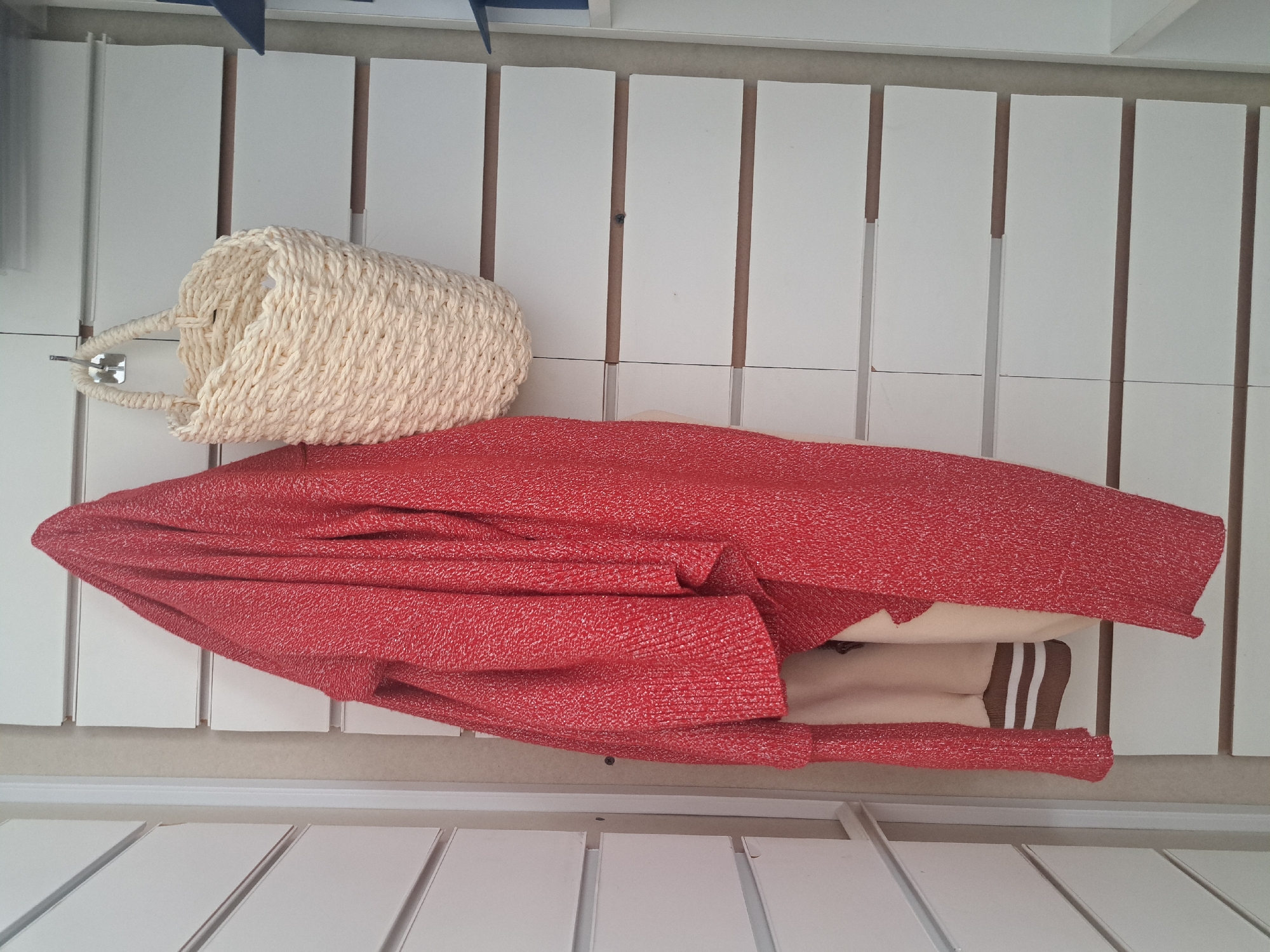 A low-angle, close-up indoor shot depicts a white wicker purse and a red knit garment resting on a white, slatted surface. Part of a tan garment with brown stripes is visible beneath the red knit. The background is composed of vertical white panels. The location is unspecified and cannot be determined from the image.