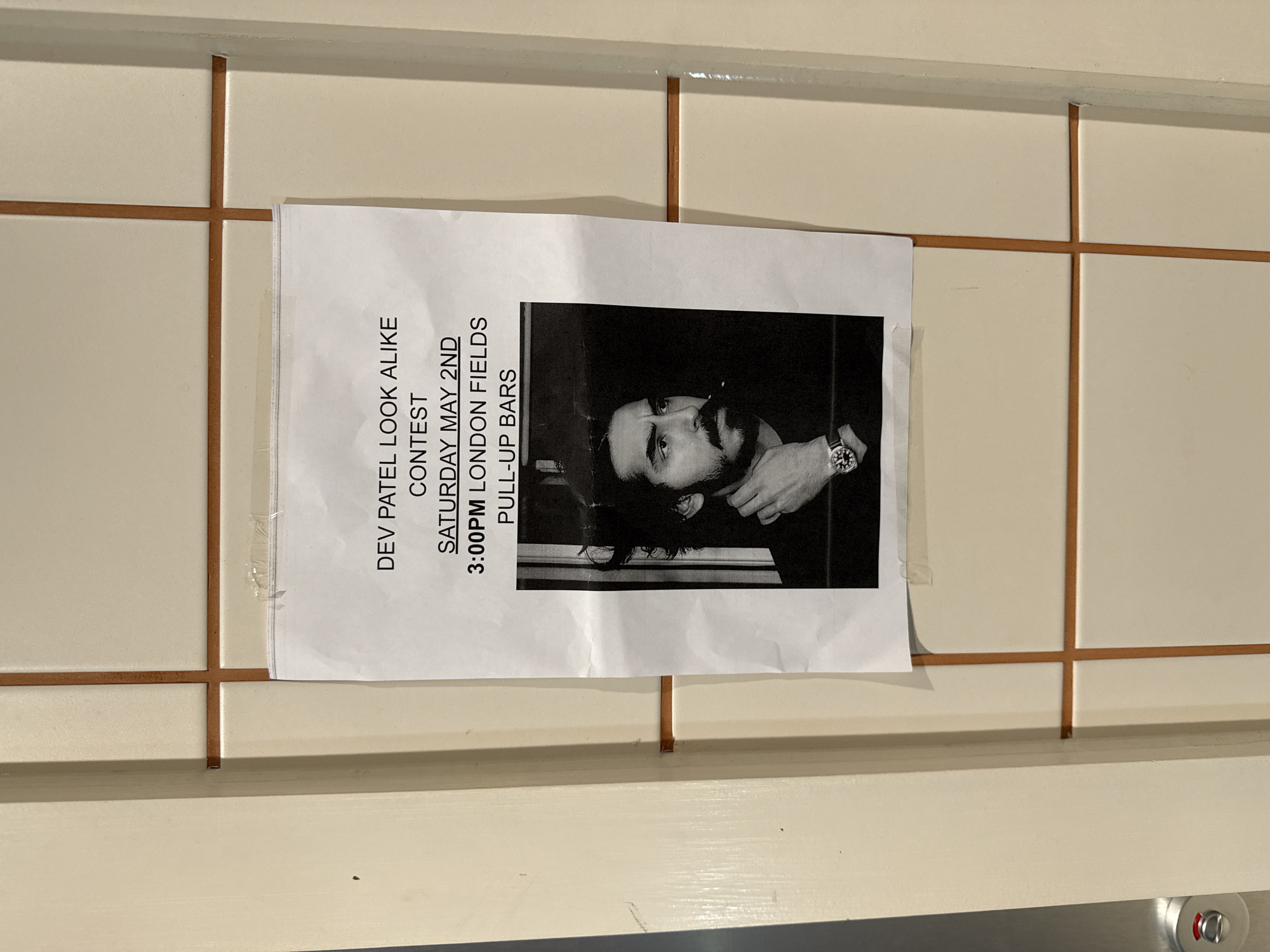 A white paper flyer, oriented vertically, is taped to a cream-colored tiled wall with brown grout lines. The flyer advertises a "DEV PATEL LOOK ALIKE CONTEST" and displays a black and white portrait of a man, presumably Dev Patel, who has dark hair, a beard, and a mustache, and is wearing a watch on his left wrist. The text on the flyer, running along its left side, provides event details: "SATURDAY MAY 2ND," "3:00PM LONDON FIELDS," and "PULL-UP BARS." The flyer is secured to the wall with clear adhesive tape visible at its top left and bottom right corners. The tiled wall features square or rectangular tiles in a grid pattern. A small portion of a silver-colored fixture with a red circular accent is visible at the bottom right corner of the overall image. The setting is likely an indoor or sheltered public space, with the event itself scheduled for London Fields, United Kingdom.