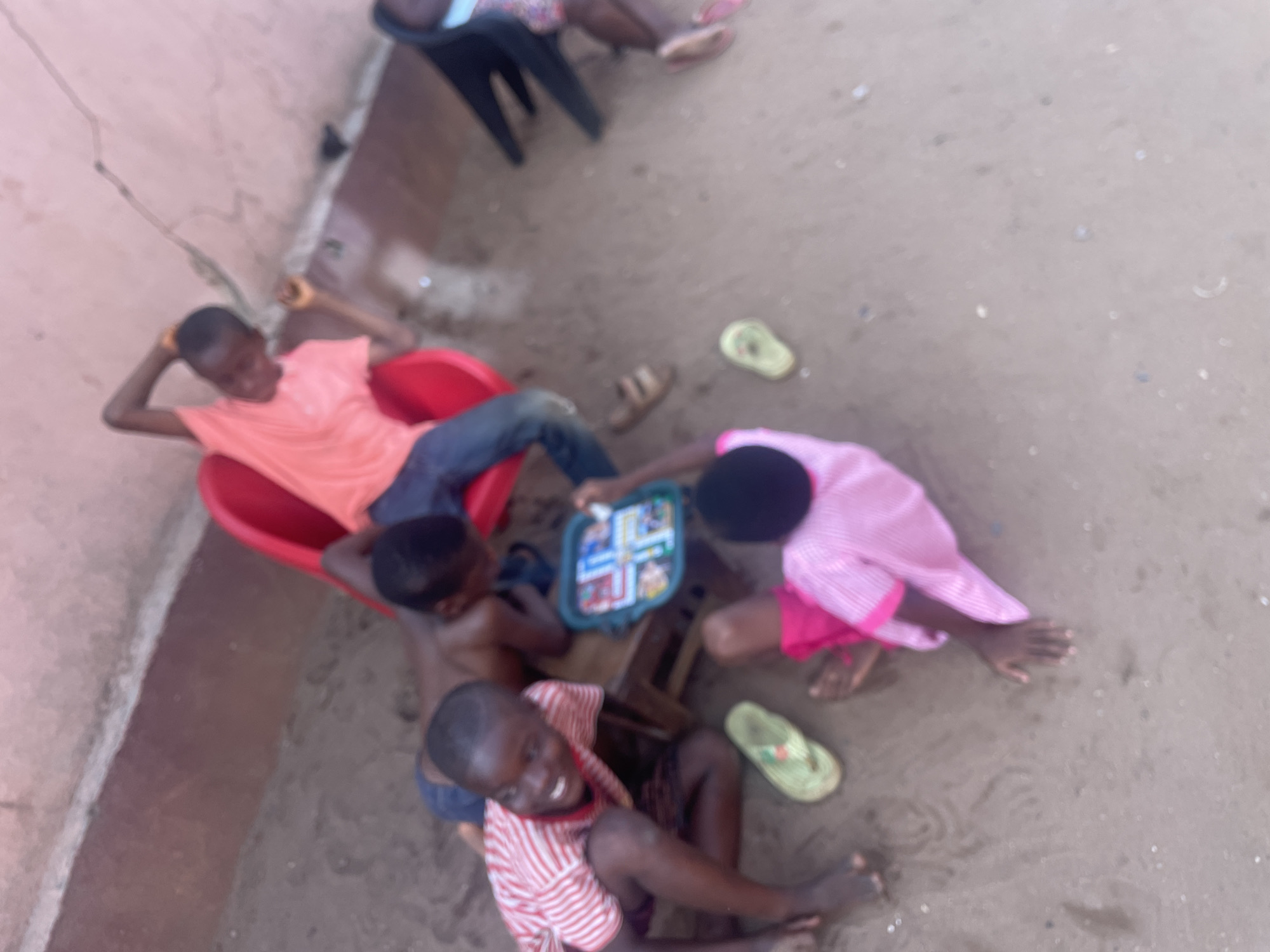 This is an overhead shot of several children playing outdoors on a sandy or dirt ground in Nkpor, Nigeria, during the daytime.
In the center-left, a boy wearing a pinkish-orange t-shirt and blue jeans is seated in a red plastic chair, appearing relaxed with his hands behind his head. Below him, another boy, shirtless and in dark shorts, is sitting on the ground, seemingly engaged in a game. To the right, a third boy, wearing a white and red striped t-shirt and dark shorts, is sitting on the ground and smiling broadly towards the camera, showing a cheerful expression.
A fourth child, dressed in a pink top, is leaning over a small wooden table, actively playing a board game that appears to be Ludo or a similar game, characterized by its colorful, segmented tracks. The board game is the focal point of the activity for some of the children. Various pairs of sandals or flip-flops, including light green and brown ones, are scattered on the ground around the playing area, indicating that the children are barefoot.
In the background, a light-colored wall with visible cracks suggests an older building structure. Parts of what appear to be adults or older children are visible in the upper left, seated on black plastic chairs. The overall scene depicts a casual, communal outdoor play setting, with the children enjoying their game under natural light. There is no discernible text visible in the image.