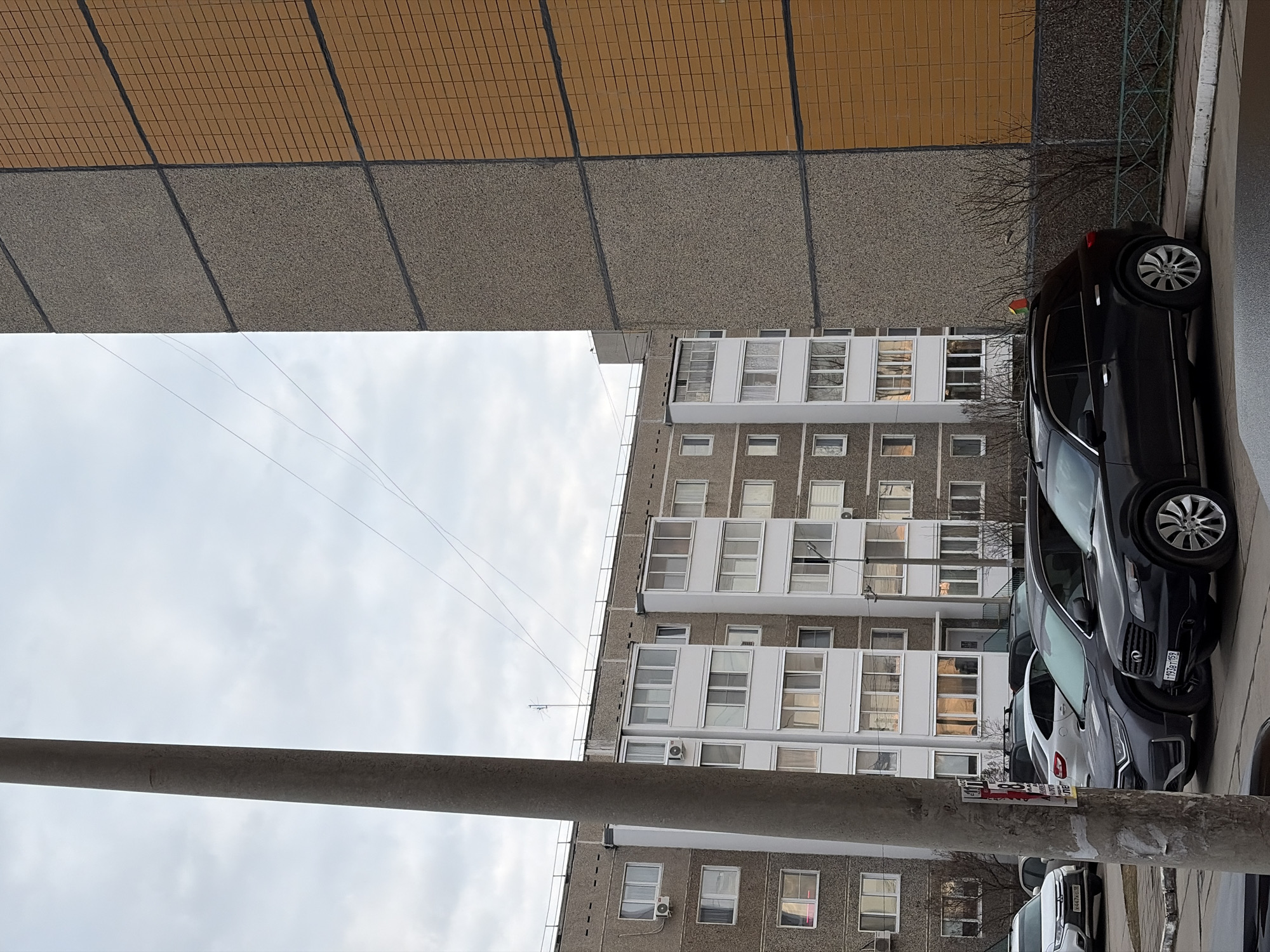 The image depicts a view in Chaikovsky, Russia, showing residential buildings, parked cars, and infrastructure. The composition is angled upwards. The buildings are multi-story, with repetitive window patterns and balconies. Several cars are parked along the street; some are black or gray. A concrete utility pole is visible in the foreground. The sky is overcast.