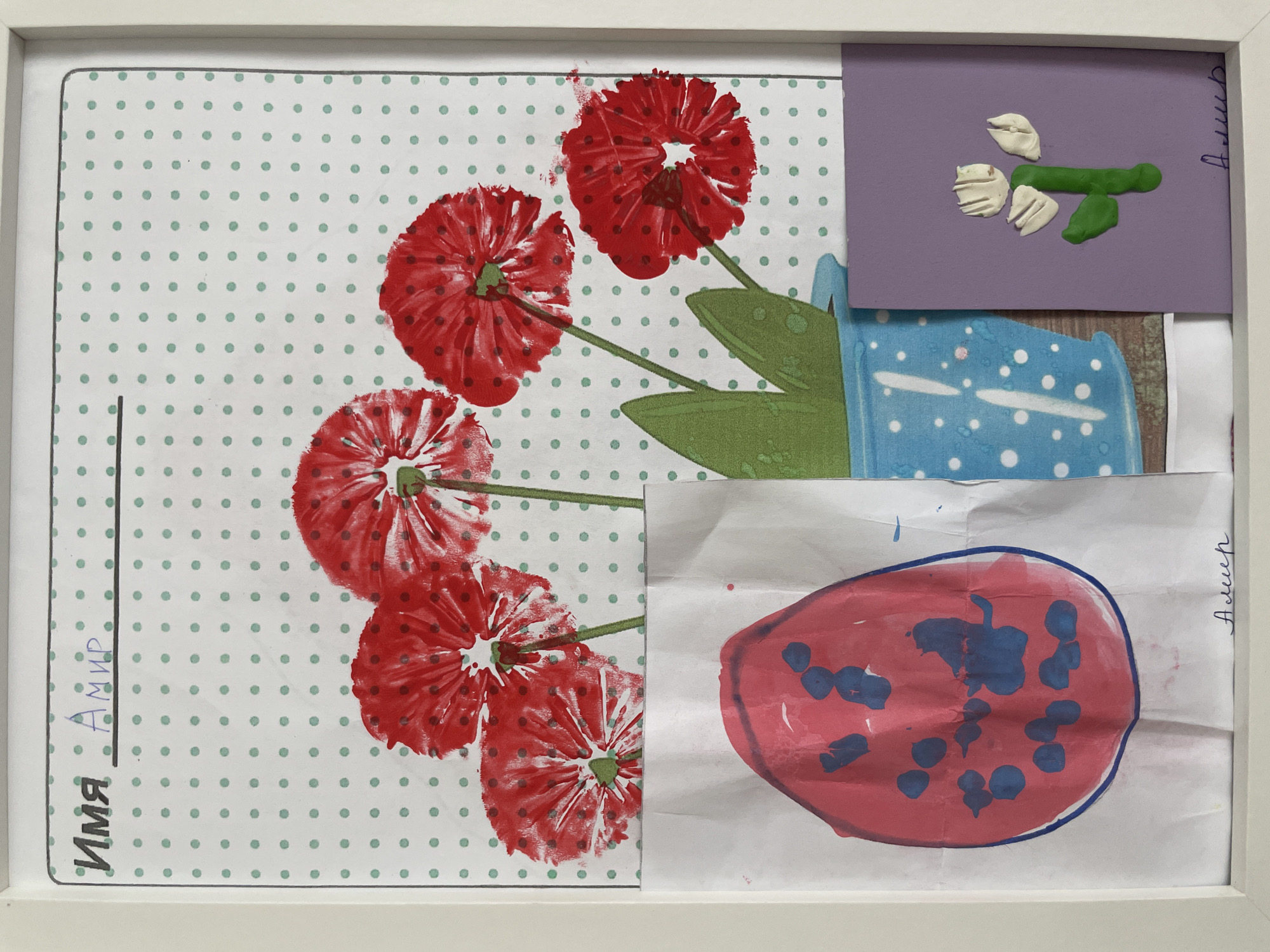 The image is of a framed collage or artwork. The frame is white. The collage consists of several different pieces of art paper or cardboard attached to a patterned background.
The background paper has a white base with a regular pattern of small, light green polka dots. On top of this background is a painting or print of red flowers resembling poppies, with green stems and leaves.
To the right of the flower artwork are several other pieces of paper. At the top is a small purple card with a green and white modeling clay decoration. Below that is a blue and white patterned piece, with a drawing of an egg-shaped form. The egg is colored with pink and blue paint.
There are handwritten words along the left side in Cyrillic letters and another word handwritten on the right side, near the egg.
There is no information regarding the location of the image in Izhevsk, Russia. The image appears to be a framed collage of artwork.