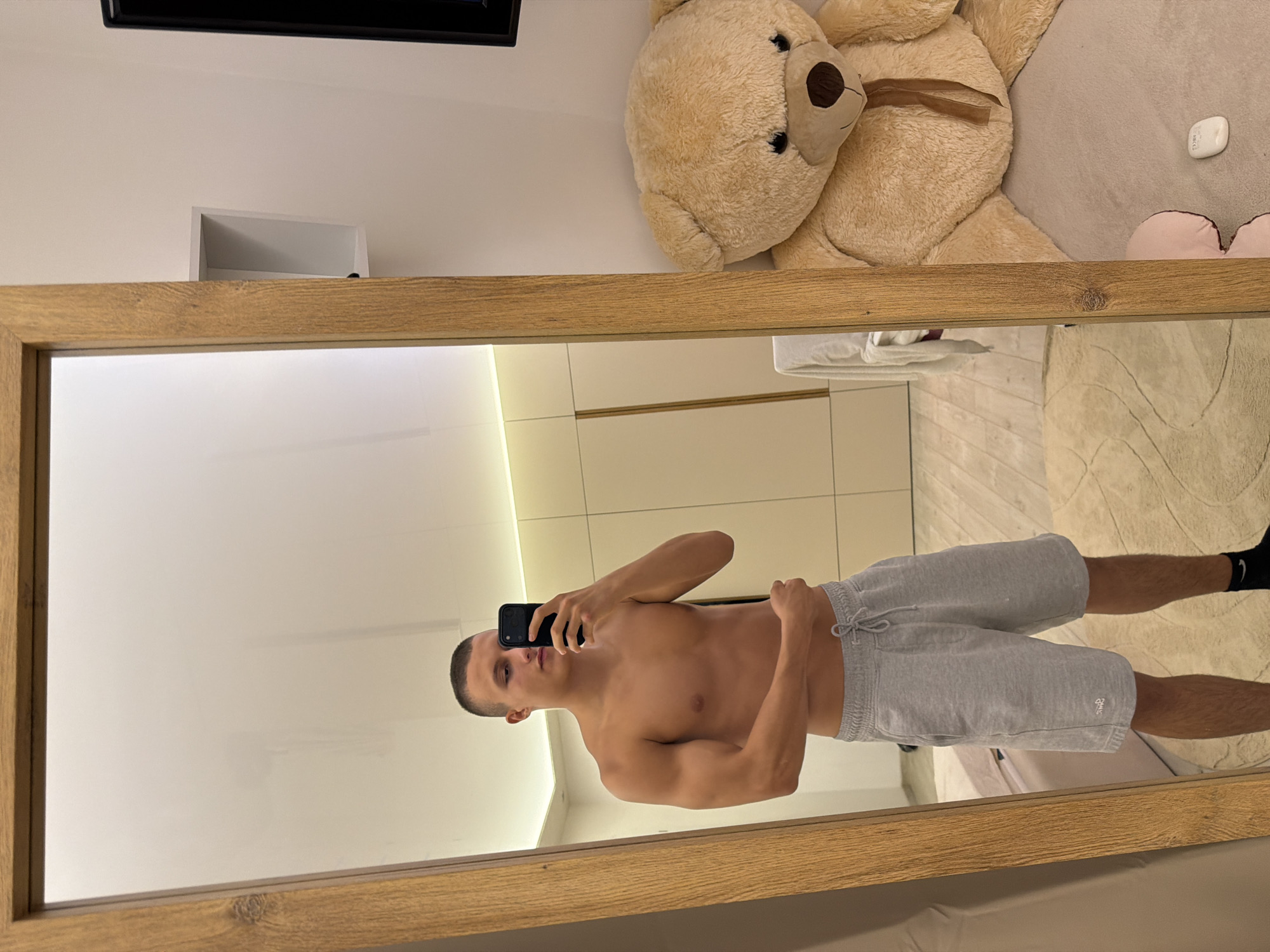 A young man with a short haircut stands shirtless in front of a full-length mirror, holding a smartphone to take a selfie. He is wearing grey sweatshorts and black socks. His physique appears athletic, with defined muscles in his arms and torso. The mirror reflects the room, which includes a large teddy bear on a bed to the right, and a wall with built-in cabinets and subtle lighting in the background. The floor is covered with a light-colored rug. The setting appears to be a bedroom or a dressing area, likely within a residential property. There is no information in the image to suggest the location is in Elhovo, Bulgaria.