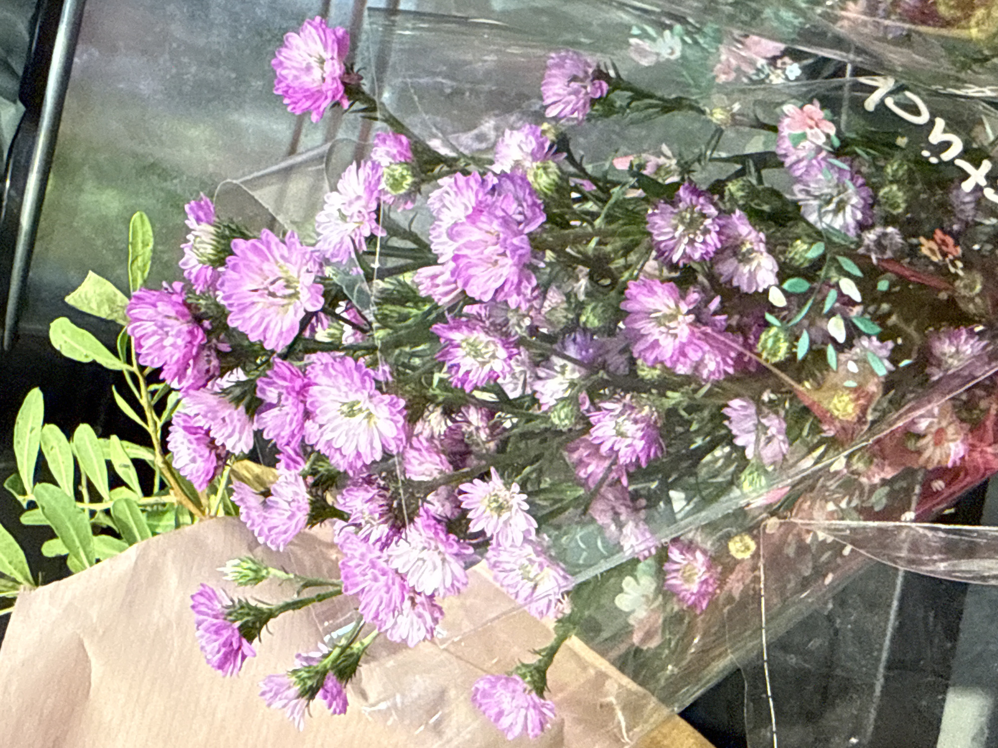 A bouquet of small, pink, aster-like flowers is displayed in clear plastic packaging. The flowers are tightly clustered and have a daisy-like appearance with numerous thin petals. Some of the flowers are fully open, revealing yellow centers, while others are still in bud form. The stems and foliage are green and appear delicate. The bouquet is partially wrapped in brown paper on the left side. The background is dark and out of focus, but a metallic surface with horizontal lines is visible at the top. Text in white is partially visible on the plastic packaging at the bottom right, reading "stück." The scene appears to be in a retail setting.