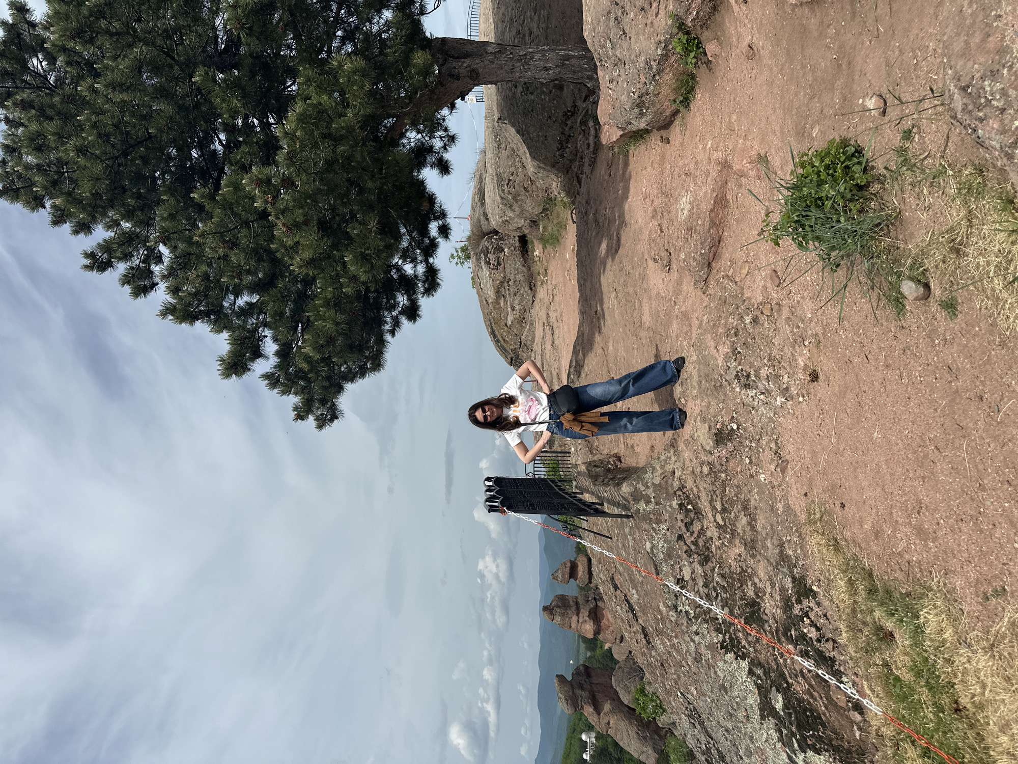 The image depicts a woman standing on a rocky, elevated terrain with a tree to her left and a panoramic view in the background. She has her hands on her hips and is wearing sunglasses, a white t-shirt, and jeans. To the right of the woman are some rocks and patches of grass. In the foreground is a safety barrier with a metal railing and a chain fence, indicating a viewpoint or a potentially hazardous edge. In the distance, the landscape consists of rocky formations, potentially the Belogradchik Rocks, and forested hills under a cloudy sky. The location is suggested to be Belogradchik, Bulgaria.