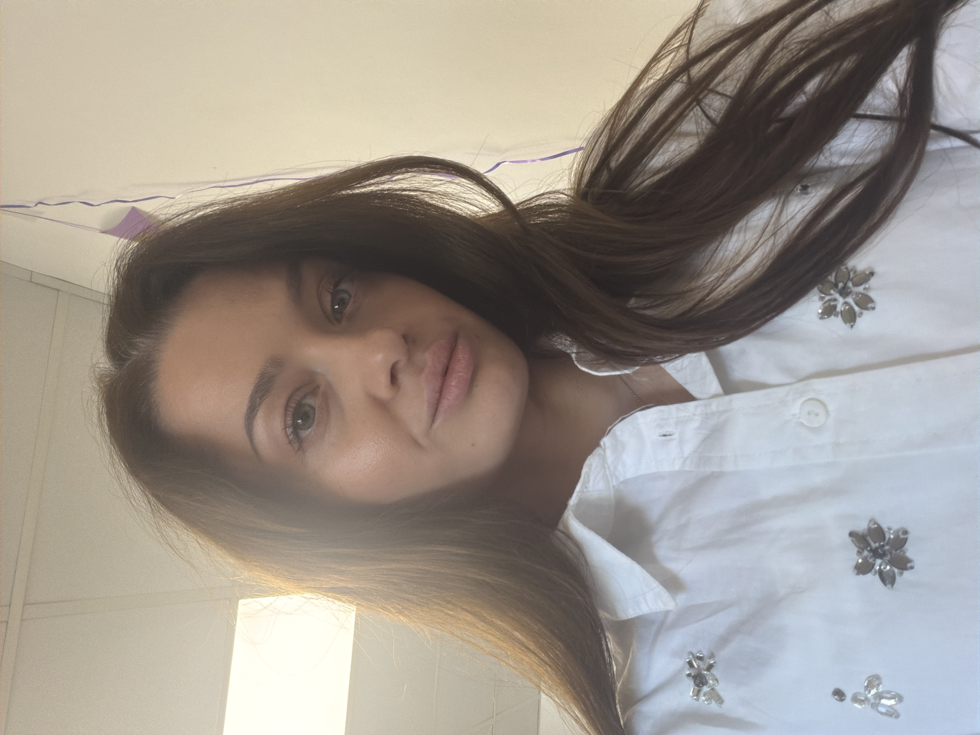 The image is a portrait of a young woman, captured from a slightly upward angle, giving the impression of a selfie. She looks directly into the camera with a serene and natural expression. Her long, straight, dark brown hair flows around her face and over her shoulders. She has full lips, appearing naturally colored or with a light gloss, and light-colored eyes, possibly green or hazel.
She is dressed in a white collared shirt, distinguished by several small, sparkling decorative embellishments on the front. These adornments resemble floral or star shapes, crafted from silver-toned stones or beads.
The setting appears to be indoors with a light-colored background, likely a wall or ceiling. A subtle, wavy purple ribbon or decoration is strung across the upper portion of the background. A bright, rectangular light source, possibly a window or a lamp, is visible towards the lower left, casting a soft, warm glow. The overall lighting is gentle, suggesting ambient daylight or soft artificial illumination. The woman is simply posing for the photograph. No text is visible within the frame. While the image is provided with the context of Varna, Bulgaria, the photograph itself does not contain any specific visual indicators of this geographical location.