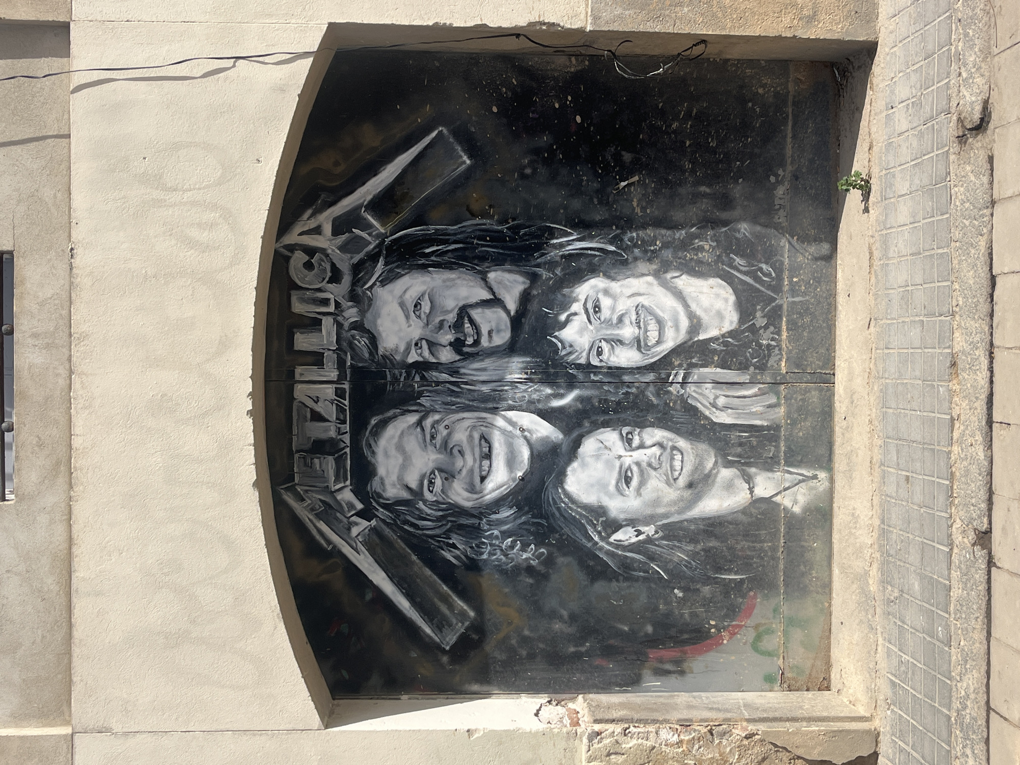 An outdoor, brightly lit image captures a street art mural depicting the rock band Metallica, painted on a dark metal door or gate. The mural, rendered in grayscale, features four caricatured faces of the band members – two on an upper section and two on a lower section – all with wide, joyful smiles and distinctive long hair. The word "METALLICA" is painted vertically in block letters along the left side of the artwork.
The mural is set within an arched opening of a building facade, which is constructed from a light-colored, textured material resembling stucco or concrete. Strong sunlight casts distinct shadows, indicating a clear, sunny day. Above the arch, a dark, thin wire runs horizontally. To the right of the arch, a section of the wall features small, square tiles in a gray tone, alongside rougher concrete elements. A small cluster of green leaves, a tiny plant, sprouts from a crack in the tiled wall, adding a touch of natural growth to the urban setting. The overall scene suggests a vibrant piece of street art integrated into a city environment, possibly in Barcelona, Spain.