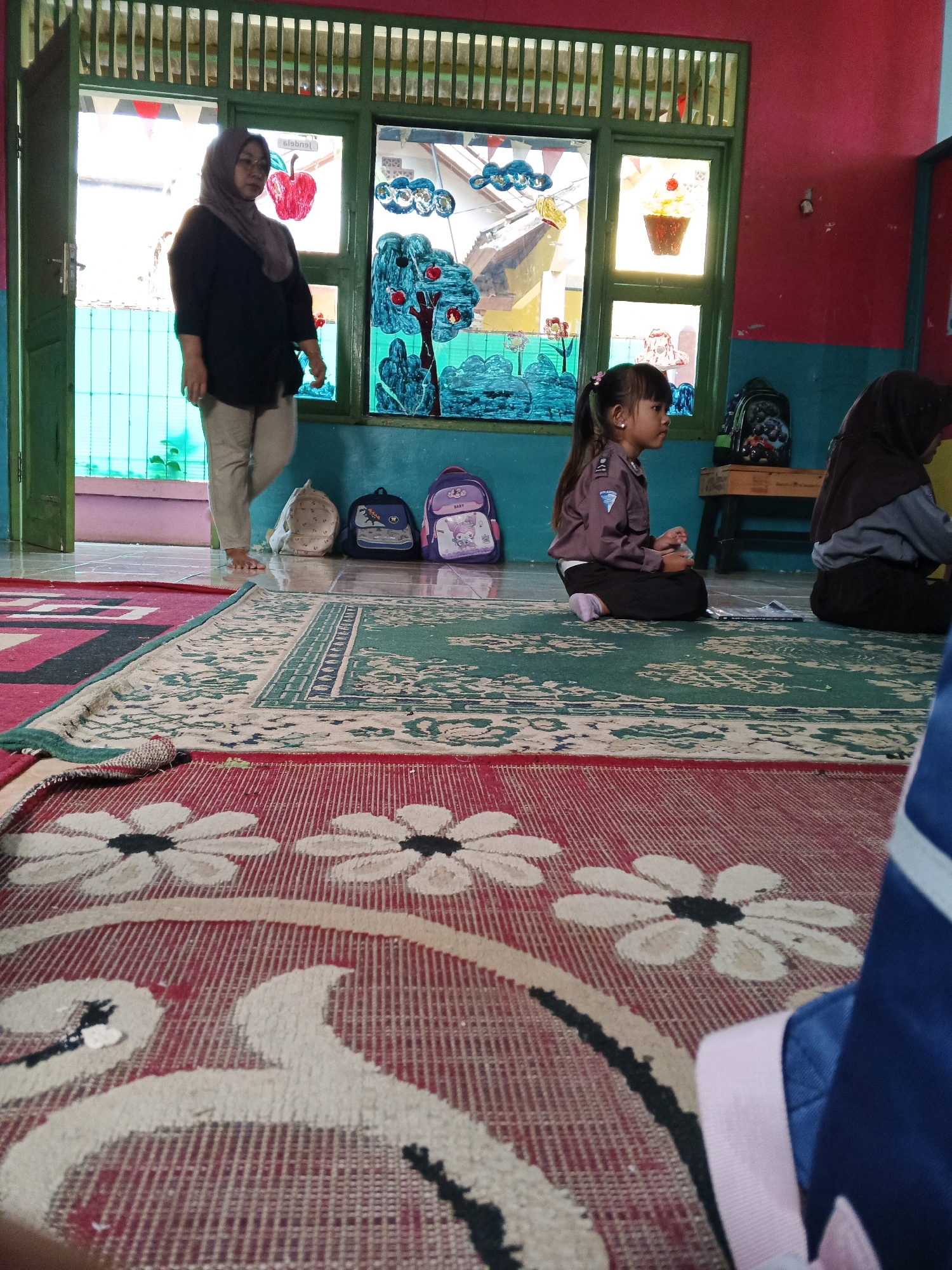 In Warungkiara, Indonesia, a classroom scene unfolds with a woman wearing a headscarf and a black shirt standing near an open doorway. A young girl in a brown uniform sits on a green carpet, facing to the right, with backpacks and a light-colored bag on the floor behind her. Another child, mostly obscured by the foreground, is also present. The room features brightly colored walls and windows decorated with cutouts of fruits and other images. The floor is covered with patterned rugs.
