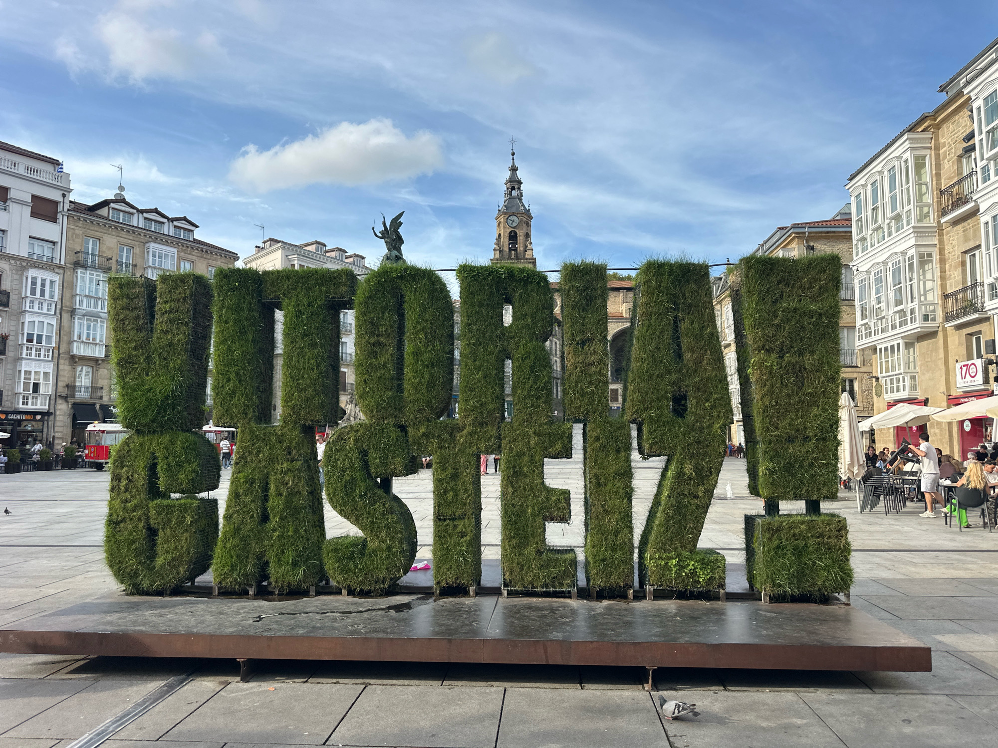 In Vitoria-Gasteiz, Spain, a large grass-covered sculpture spells out "VITORIA-GASTEIZ" in a public square. The sculpture consists of individual, towering letters, each made of dense, green grass and supported by a dark, metallic base. The letters are spaced apart, creating gaps between them, allowing the background to be visible.
Behind the sculpture, a street stretches out, lined with buildings. The architecture features a mix of styles, including buildings with stone facades, ornate balconies, and large bay windows. A prominent clock tower with a spire rises above the buildings in the distance, its face visible. The sky is a bright blue with scattered white clouds.
In the mid-ground, people are present in the square. Some are seated at tables outside a cafe, enjoying drinks and conversation under umbrellas. Others are walking or standing, some interacting with the sculpture. A red tram-like vehicle is visible in the distance, on what appears to be a tram line. A statue of a winged figure is perched on top of one of the grass letters. Pigeons are seen on the paved ground in the foreground and mid-ground. The overall scene depicts a lively urban public space on a clear day.