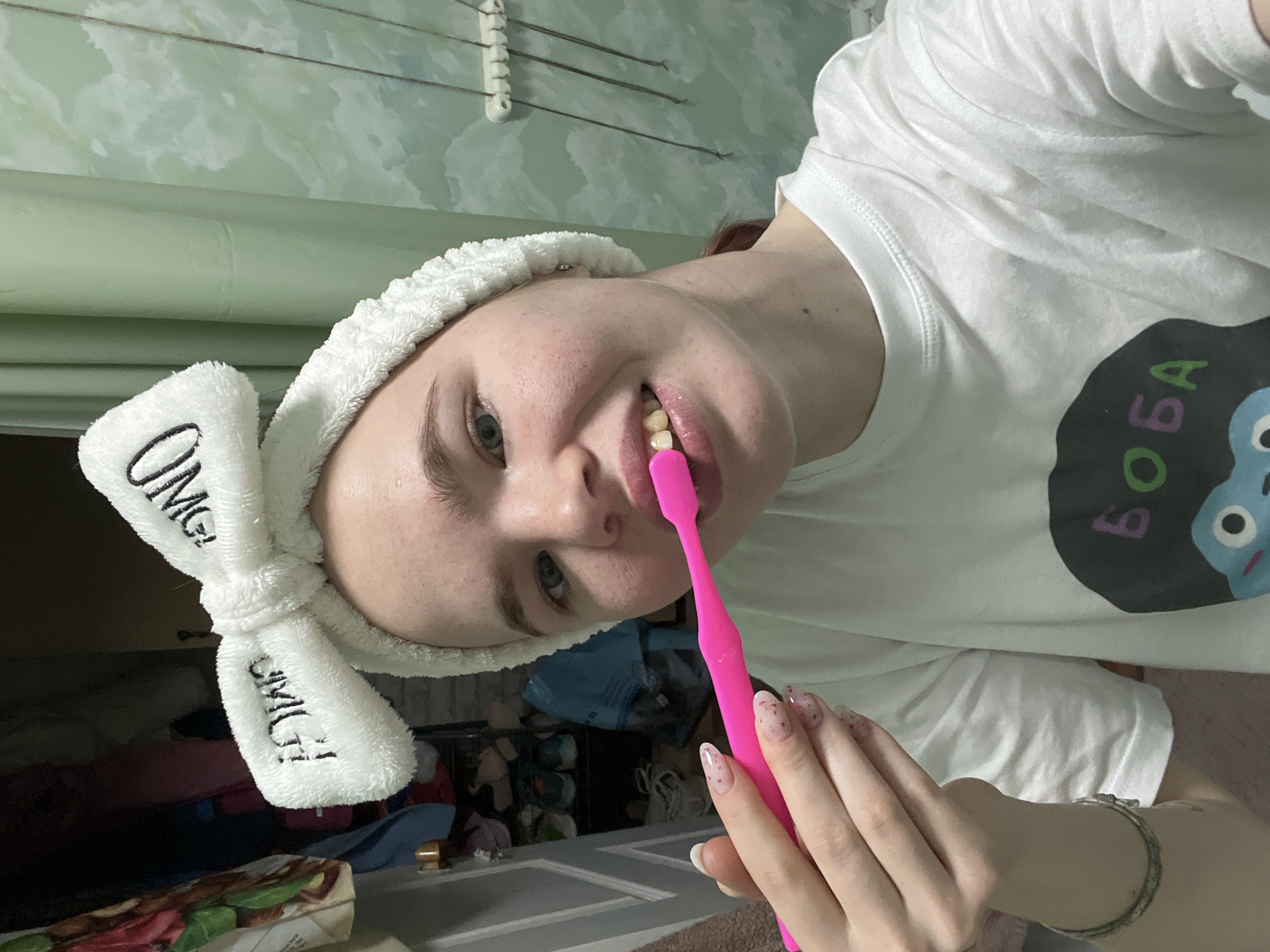 A young woman is seen in an indoor setting, likely a bathroom, given the green painted wall and towel rack in the background. She is angled towards the camera, wearing a white t-shirt with a large, cartoonish design on the front. She is also wearing a fluffy white headband with the word "OMG!" written on it. She is in the act of brushing her teeth with a pink toothbrush. Her nails are manicured with a light pink base and small, scattered red and white heart-shaped accents. The background is somewhat cluttered, with items on a shelf or in a cabinet, suggesting a domestic setting. It is not possible to determine the exact location in the city of Chaikovsky, Russia, without additional information.