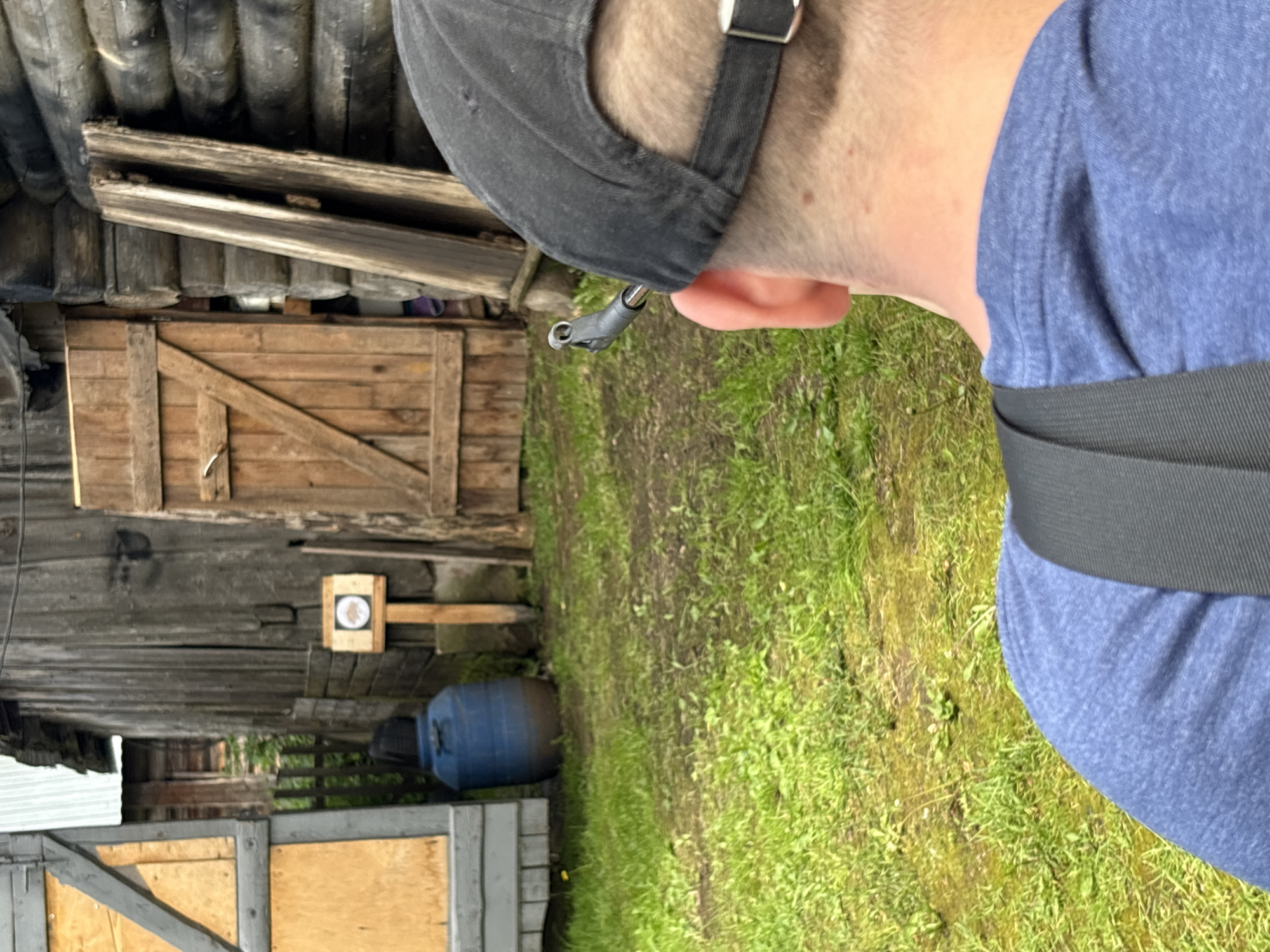 A person wearing a blue t-shirt and a black baseball cap is viewed from behind in the foreground. The cap has a black strap with a buckle at the back. Attached to the rear of the cap is a small, dark, L-shaped device. The person appears to be looking towards a wooden structure and a target. In the background, there is a weathered wooden building with a door and a target mounted on a wooden post. The target is round and has a picture of an animal inside a circle. To the left of the building, there is a blue plastic barrel and part of another wooden structure. The ground is covered with green grass and dirt. The scene appears to be outdoors in a rural setting, possibly in the village of Дресвяново, Russia.
