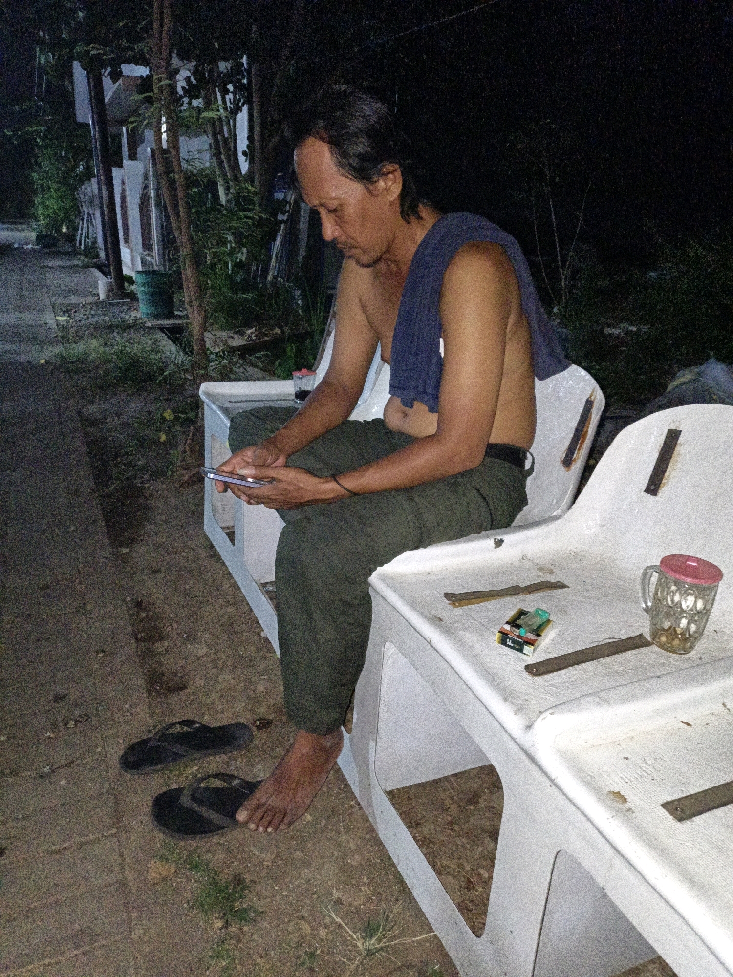 A man is sitting on a white plastic bench in what appears to be an outdoor setting at night. He is holding a smartphone and looking down at it. He is shirtless, with a dark blue cloth draped over his right shoulder. He is wearing dark green pants and black flip-flops are on the ground next to him. On the bench in front of him are a pack of cigarettes, a lighter, and a glass mug with a pink lid. The bench is situated on a dirt and gravel area, with some vegetation and trees visible in the background. The scene suggests a casual, possibly residential, outdoor area in Cikal, Indonesia, likely a backyard or a communal seating area.