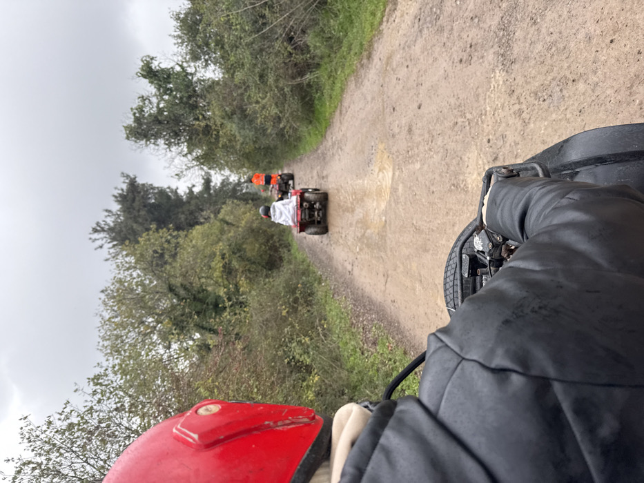 The image presents a first-person view from a quad bike navigating a muddy dirt track in Çiftlikköy, Turkey. In the immediate foreground, the left shoulder and arm of an individual wearing a black jacket are visible, along with a portion of a red helmet on the left side. The individual's hands are gripping the handlebars of a black quad bike. Ahead, two additional quad bikes proceed in a single file along the unpaved track, which features multiple visible puddles. The first quad bike in the line is red, driven by a person wearing a light-colored hooded garment and a white helmet. The second quad bike is dark-colored, operated by an individual dressed in an orange high-visibility jacket and a dark helmet. The track is bordered on both sides by dense vegetation, consisting of trees and shrubs with green and brown foliage. The sky above is uniformly overcast and grey.