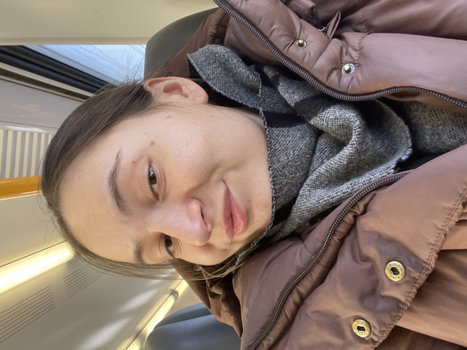 A young adult female is depicted in a close-up, selfie-style photograph taken inside what appears to be a public transport vehicle, specifically a train or subway car, in Метрополитеновец Снт, Russia. The subject has dark brown hair, parted on the side, and is looking directly at the camera with a subtle, closed-mouth smile. Her skin is fair, with a few small moles visible on her right cheek. She is wearing a dark mauve or brown puffer jacket with a visible front zipper and two golden snap buttons on the lapel area. Around her neck is a grey, possibly patterned, scarf. The background shows the interior of the vehicle, including white overhead fluorescent lighting fixtures, a yellow/orange handrail or strap towards the upper right, and a glimpse of a window revealing a bright, possibly overcast, exterior on the right side. A dark seat is partially visible behind her to the left. The scene is illuminated by interior lighting and natural light from the window.