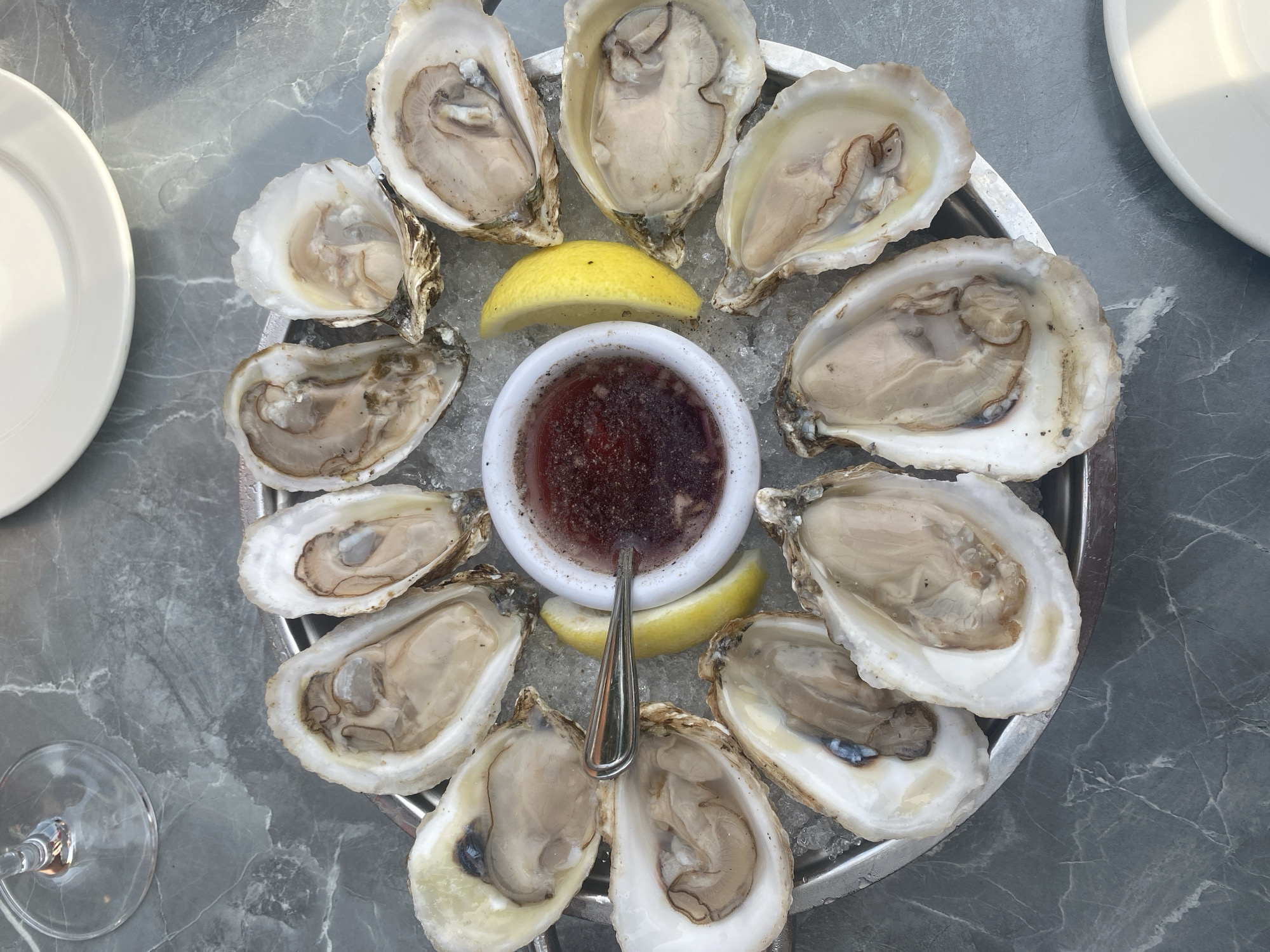 The image shows a platter of raw oysters on ice, a small white bowl of dipping sauce, and two lemon wedges, arranged on a gray, marble-patterned surface. A portion of a clear wine glass is visible in the upper left corner, and a white plate is partially visible in the upper right. The oysters are served open in their shells, revealing the glistening meat. The dipping sauce appears to be a vinaigrette or mignonette, with visible specks of herbs or spices. The setting is likely an outdoor dining area, suggested by the natural light and the marble-like table. No people are visible in the image. The overall impression is one of a fresh and appetizing seafood presentation. The context of Montreal, Canada, is not discernible from the image itself.
