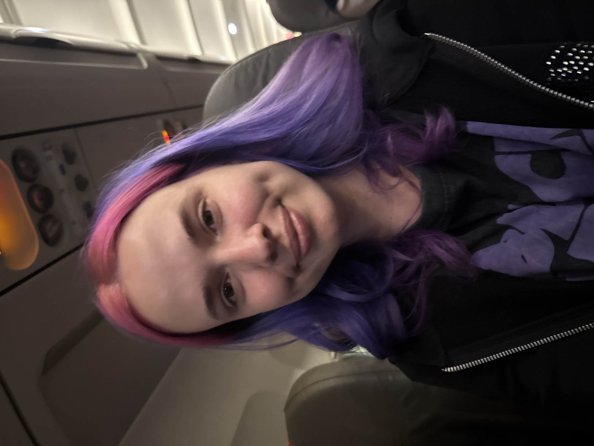 A young woman with pink and purple hair is seated in an airplane seat. She is wearing a black jacket with a zipper, and a dark t-shirt with purple graphics. The background shows an airplane interior, including the side panel with buttons and the window. The location is not visible.