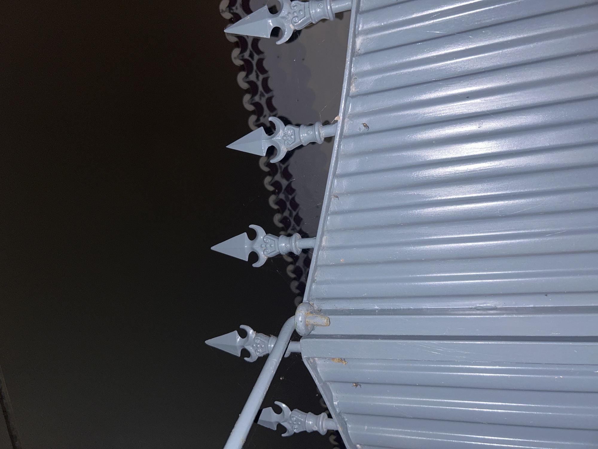 The image shows a close-up of a gray metal gate or fence. The gate has horizontal slats or panels. Projecting from the side of the gate are decorative spear-like finials mounted on metal rods. In the background is a dark, reflective surface, possibly a polished floor. The environment suggests a location in Câmara de Lobos, Portugal, characterized by architectural details often seen in the region, although the image lacks specific details to confirm the location.
