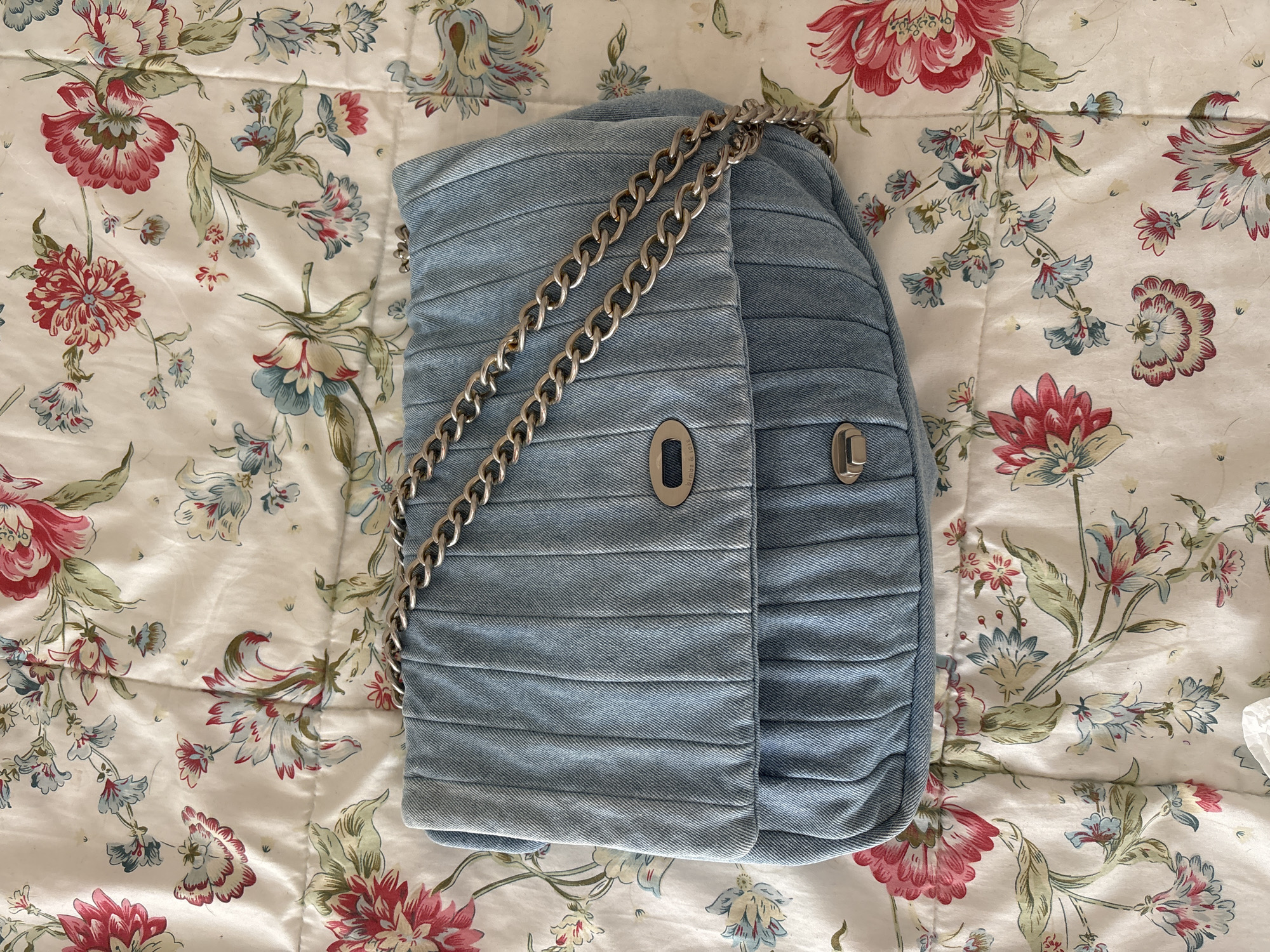 A light blue denim purse with a quilted pattern and a silver chain strap lies on a white fabric with a floral print. The purse has a flap closure with a silver metal clasp and two silver snap closures on the front pocket. The floral print consists of pink, red, and blue flowers with green leaves. The image appears to be taken indoors, possibly in a bedroom or living room, given the presence of the fabric. The image does not provide any information about a location in Orihuela, Spain.