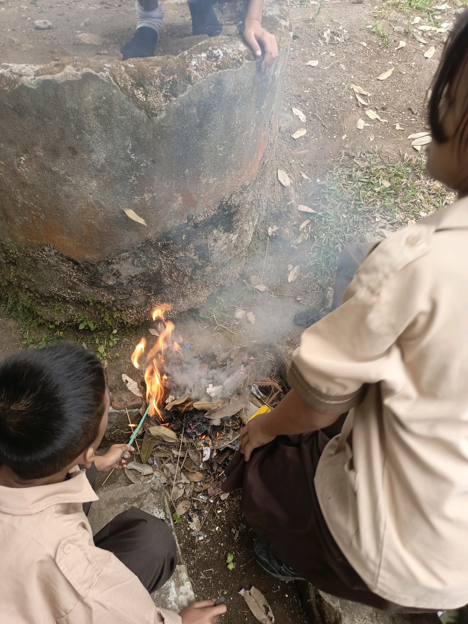 Two children, dressed in light brown uniforms with dark brown pants and white socks, are tending to a small fire. The fire is burning leaves and other debris in a pile on the ground near a large, rough concrete structure. The concrete structure appears to be a well or some kind of circular containment. The ground around the fire is dirt and dry leaves, with some sparse green grass growing. The child on the left is using a green stick to poke at the fire. The child on the right is kneeling and partially obscured by the fire, with their hand resting on the ground. The scene suggests an outdoor environment, possibly a schoolyard or a park. The lighting indicates it is daytime.