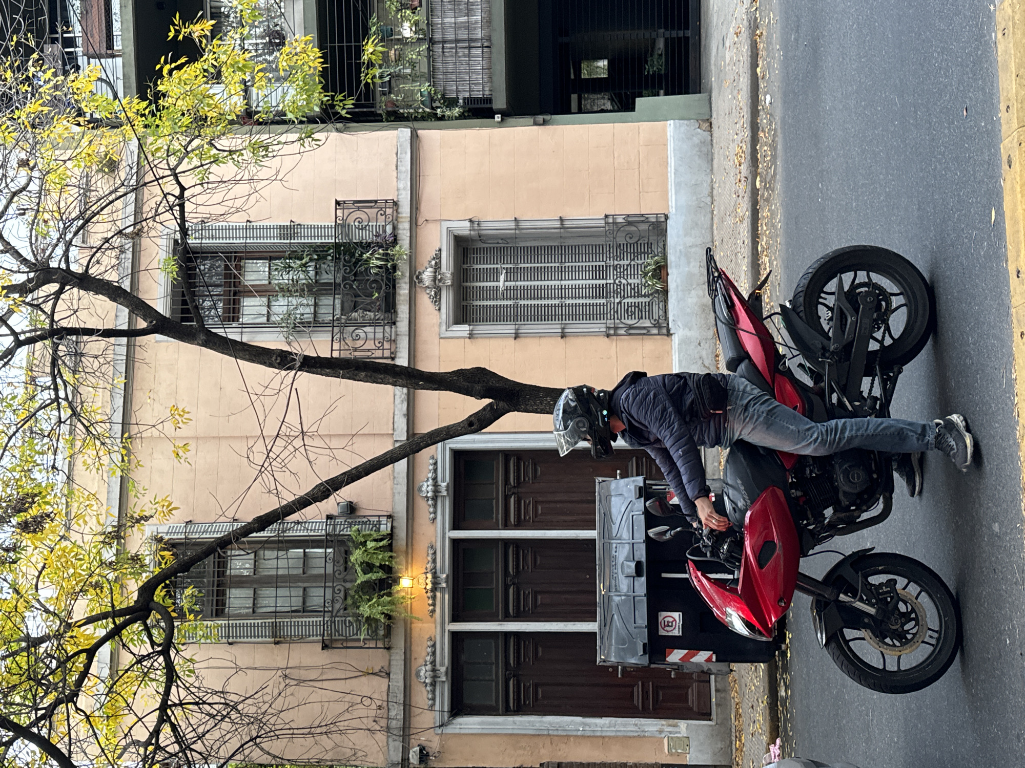 A man wearing a helmet and dark jacket is sitting on a red motorcycle parked on the right side of a street. The motorcycle has a large, dark gray container attached to the back, labeled with a red and white "no parking" symbol. The background features a three-story building with a pale peach facade, ornate ironwork on the windows, and a tree with yellowing leaves in the foreground. The location is likely in Buenos Aires, Argentina, based on the architectural style and the "no parking" symbol seen in Argentina. The street appears to be a one-way street, with no other visible traffic.