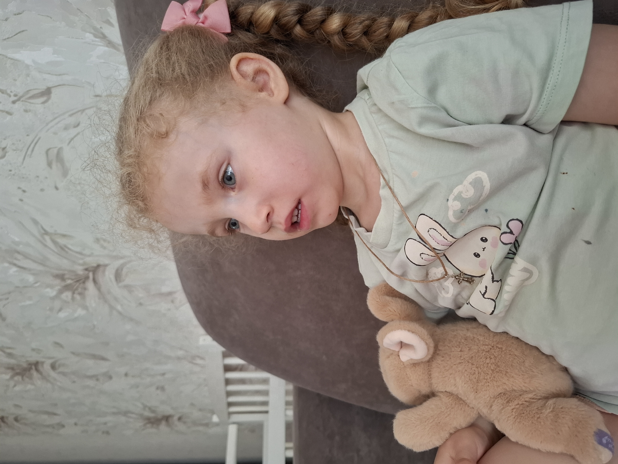 A fair-skinned, blonde-haired child with blue eyes, wearing a light green t-shirt with a bunny graphic and a thin necklace with a cross pendant, is lying down, cradling a stuffed bunny toy. The child has her hair styled in braids with a pink bow. She is resting on a brown upholstered surface against a background of white and gray patterned wall. The location is within the city of Chaikovsky, Russia.