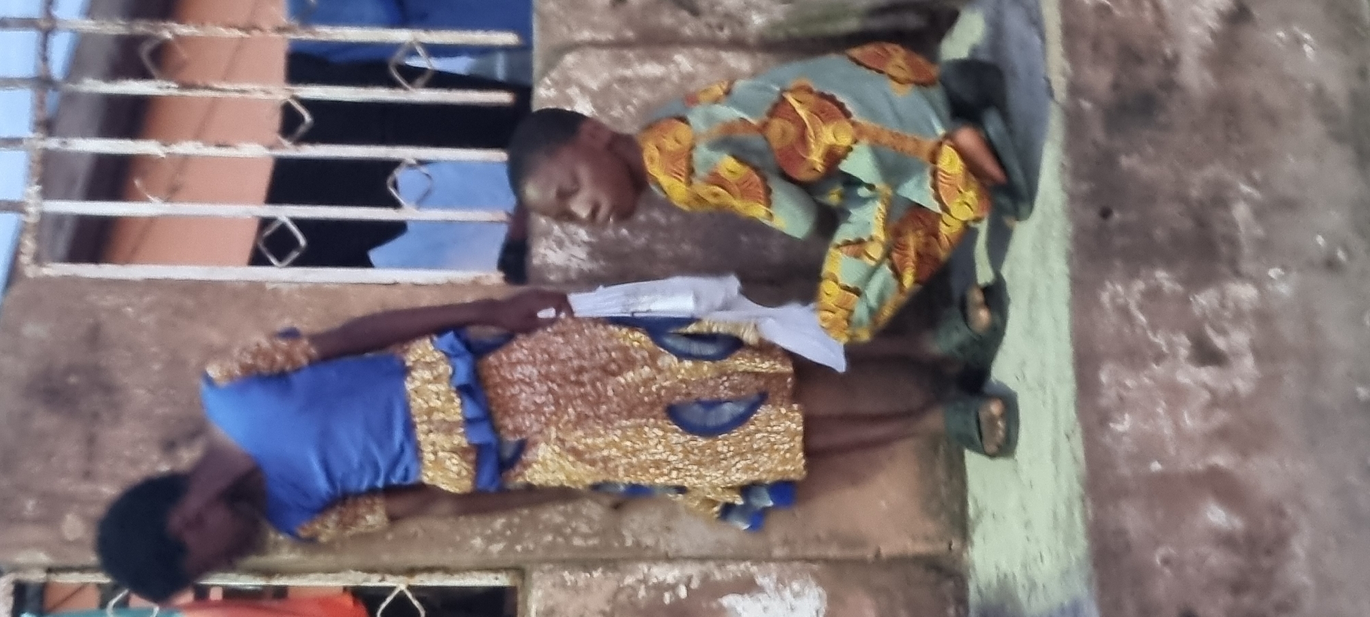 Two individuals are positioned next to a building with a barred window. One person, wearing a blue top with gold embellishments, is holding white papers. The other individual wears a light green and orange patterned top and is bent over. Both individuals are wearing sandals. The background appears to be a concrete exterior wall. The location is in an urban setting, specifically Lagos, Nigeria.